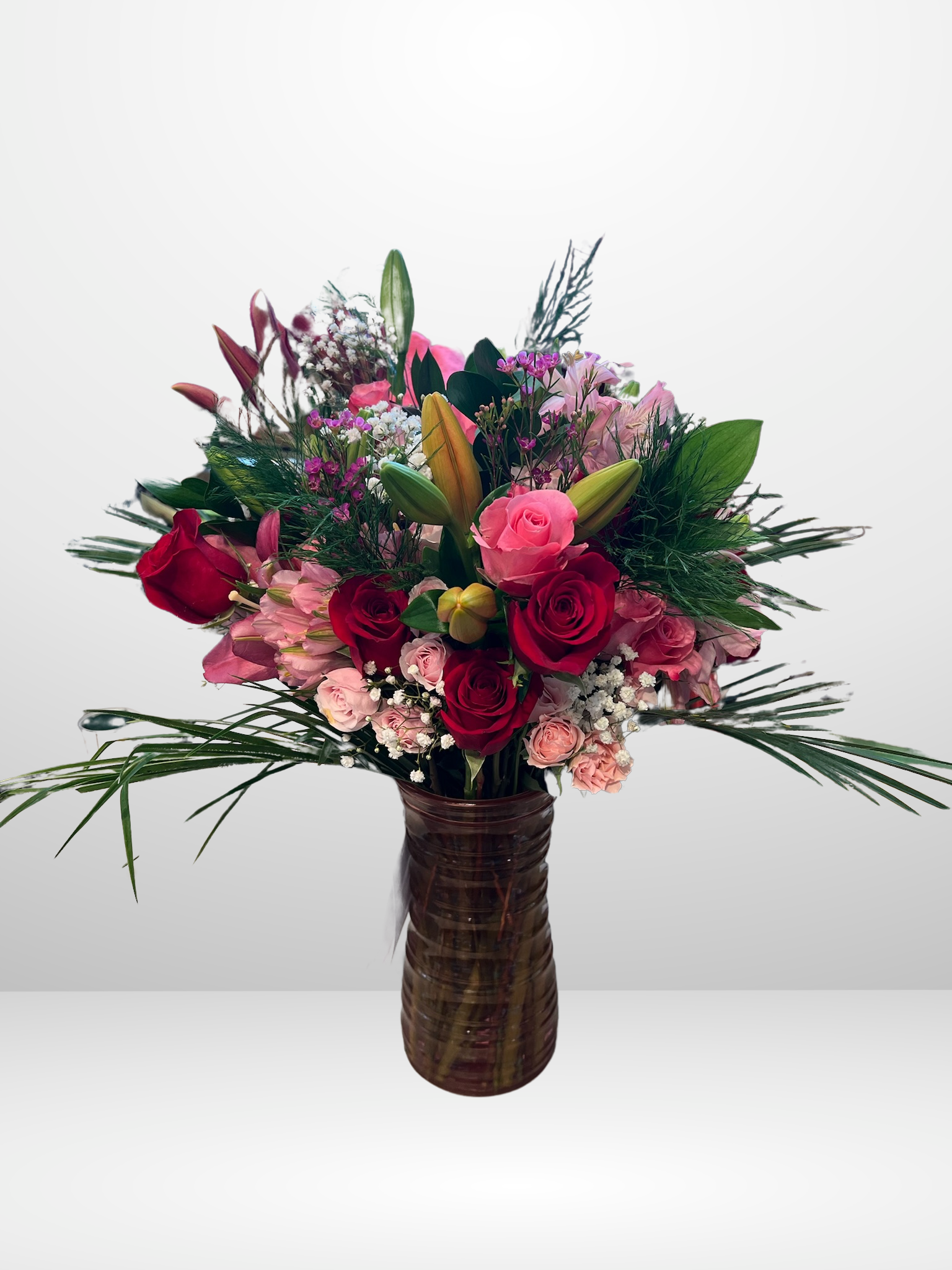 Bouquet of assorted flowers in vase with red roses, pink roses, lilies, and greenery on white background.