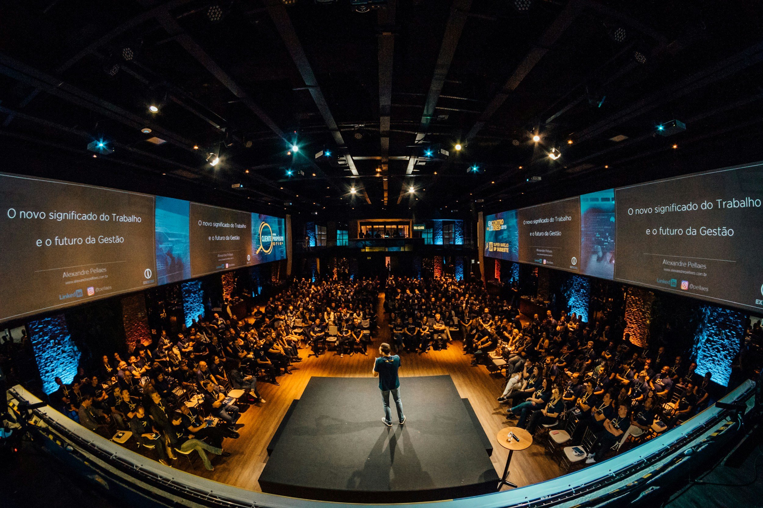 A speaker on stage at a conference, presenting to a large audience in a dark auditorium. Large screens display presentation slides with text in Portuguese.