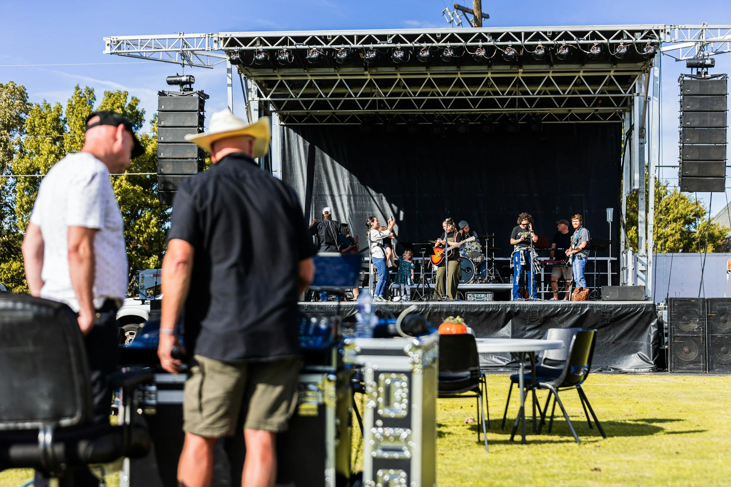 A small outdoor music concert on a sunny day with a band on stage and some people in the audience near sound equipment.