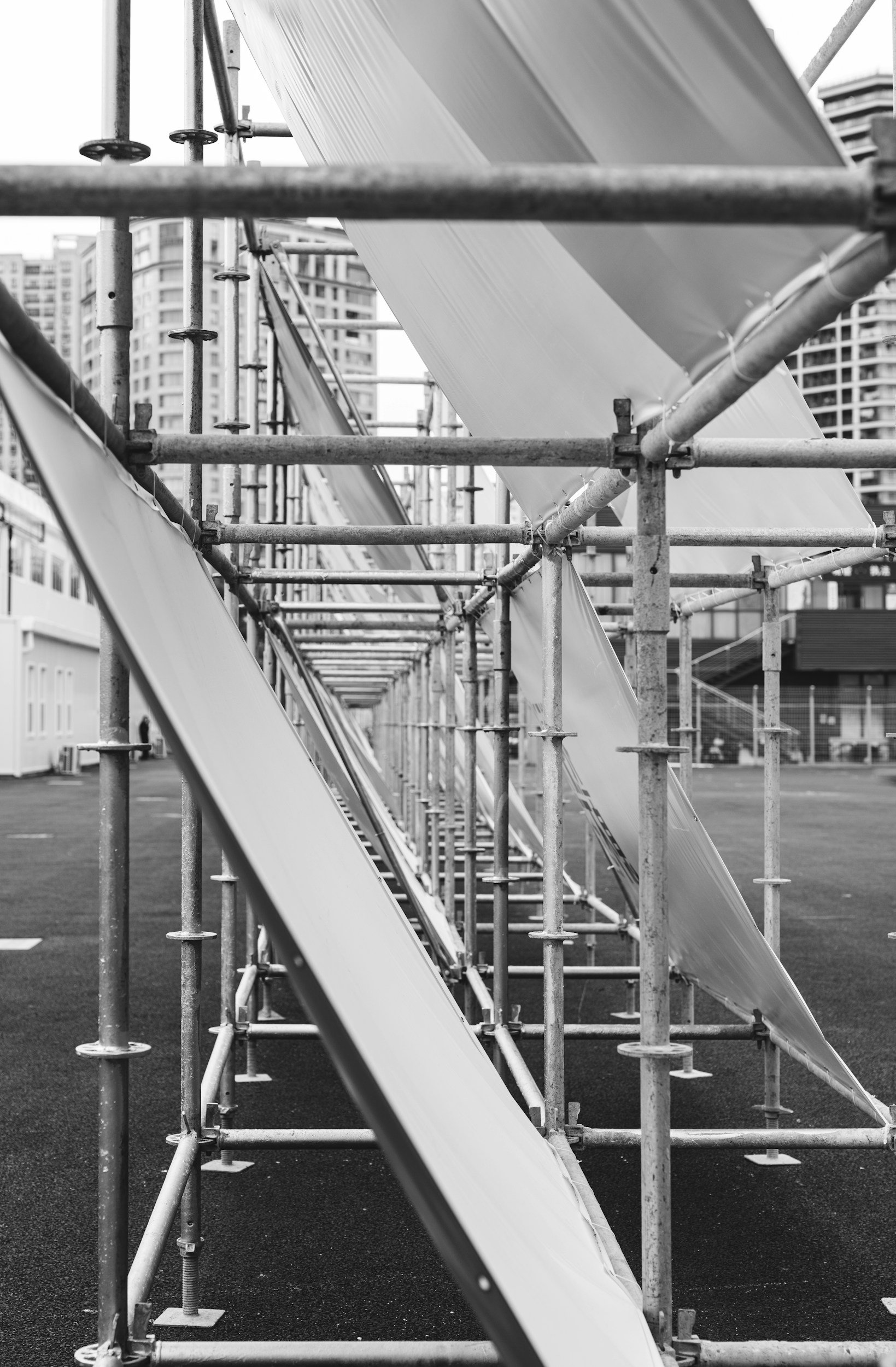 Black and white photo of a multi-story building structure under construction with metal scaffolding and plastic sheeting, with tall city buildings in the background.