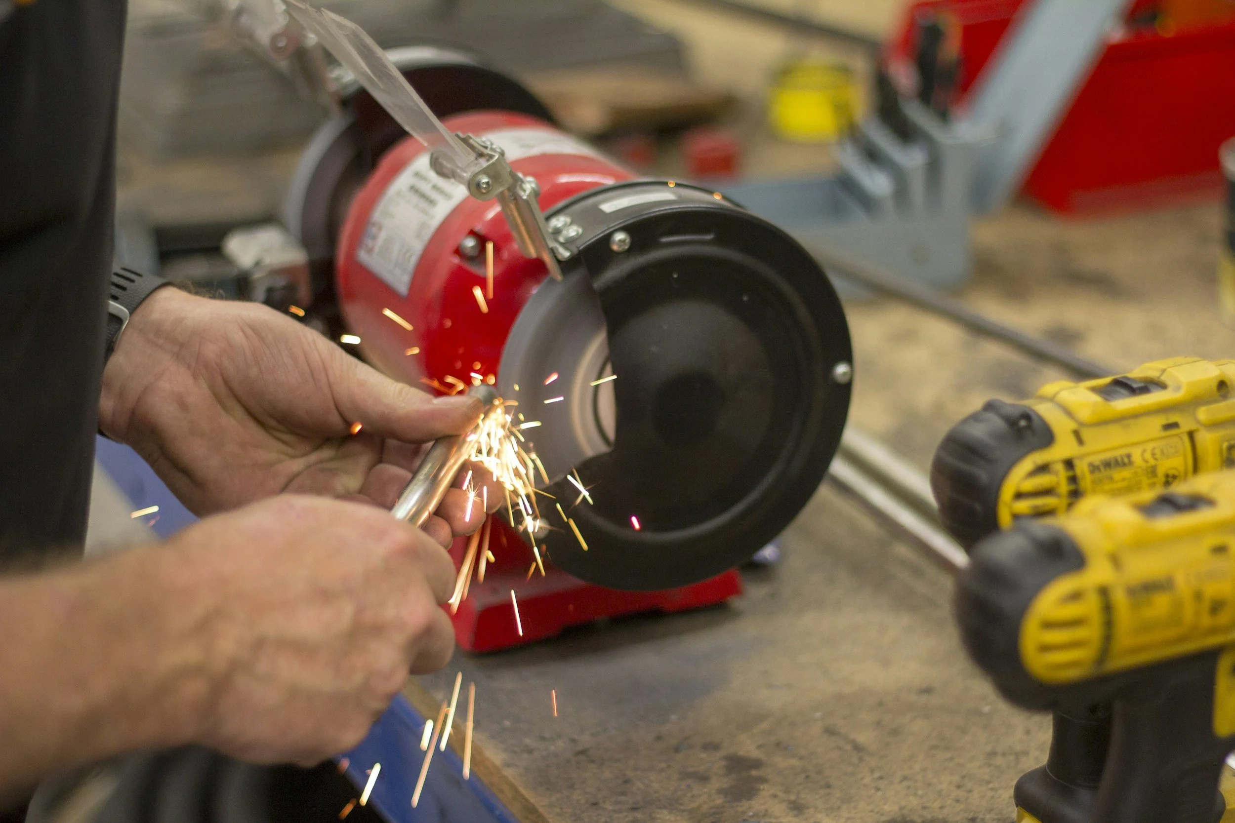 A person using a welding tool to cut a metal pipe in a workshop with a metal cutting saw and yellow power drills on the workbench.