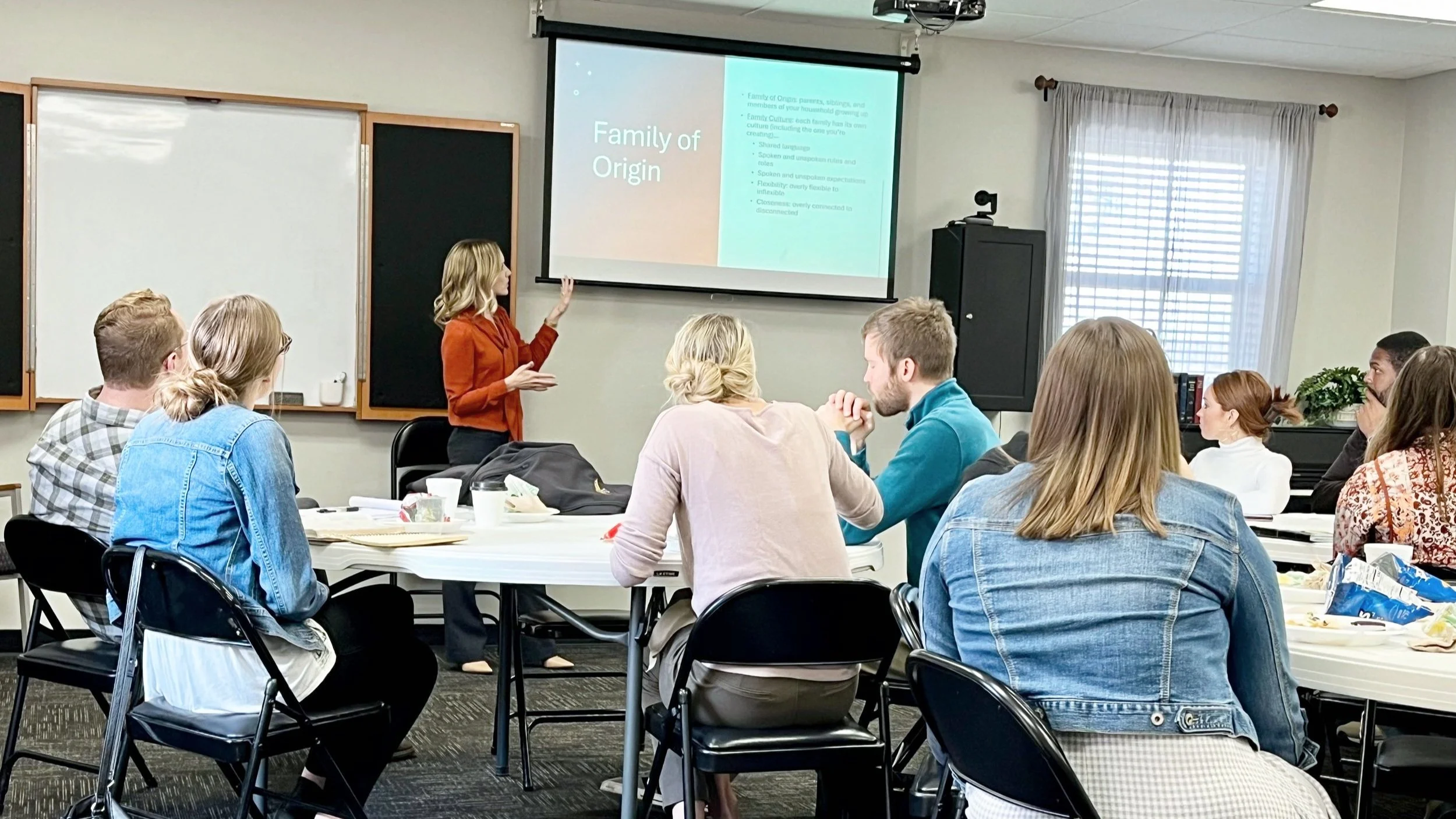 Kensi Duszynski, wearing a burnt orange blouse, giving a presentation on 'Family of Origin' to a group of people sitting at tables in a classroom setting.