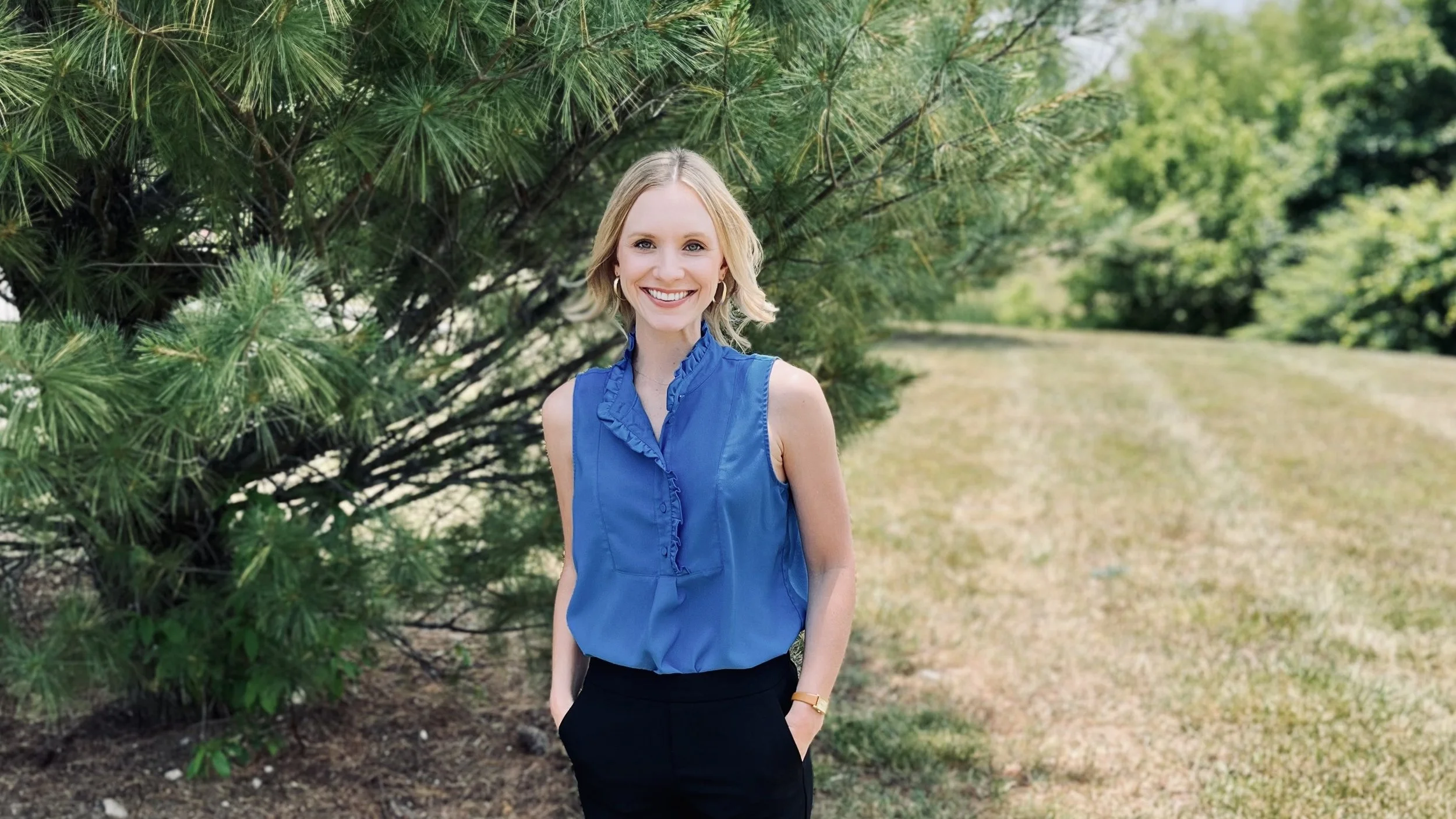 Kensi Duszynski in blue top and black pants standing outdoors near a tree, smiling.