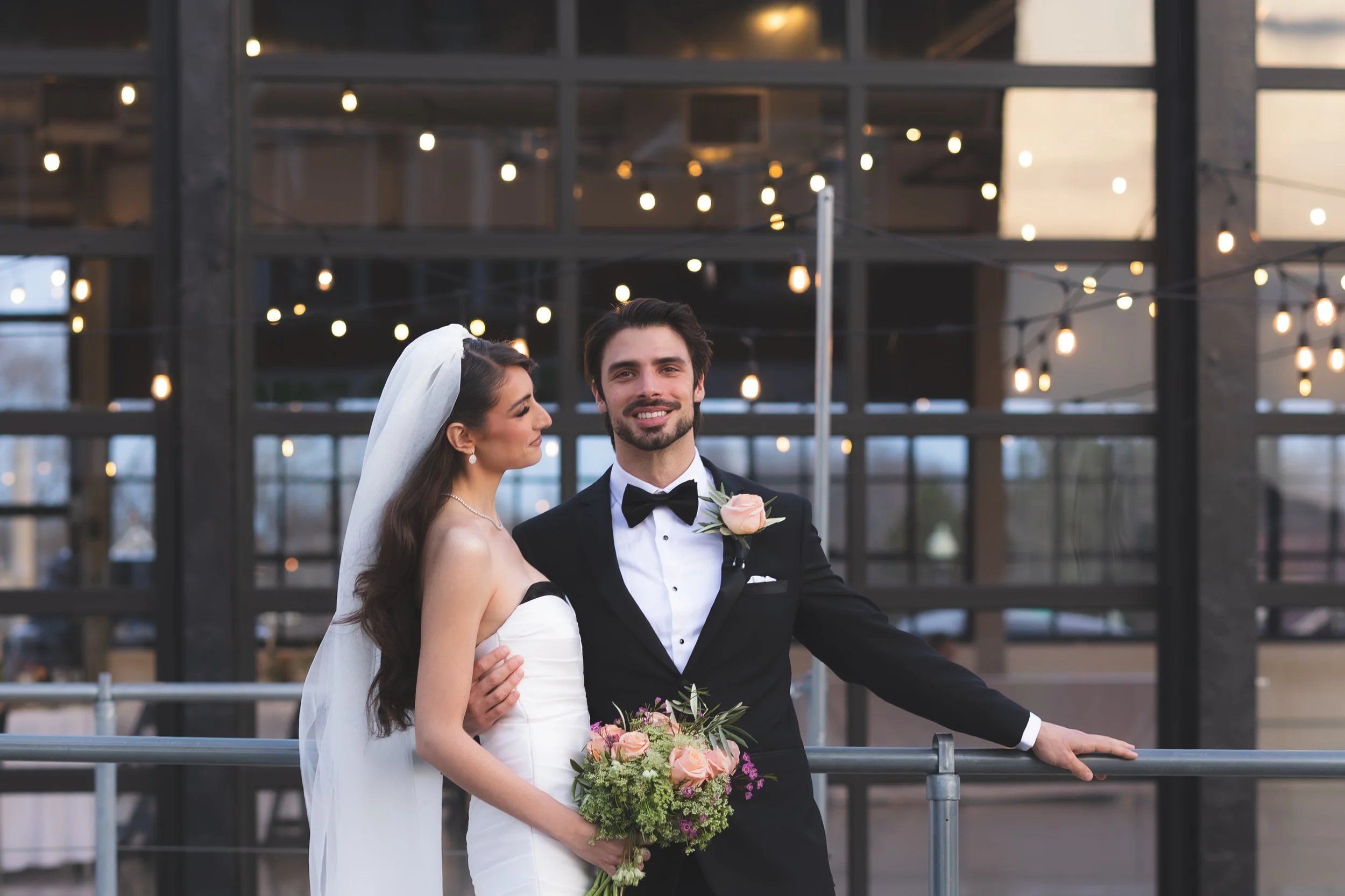 A bride and groom on their wedding day, standing outdoors at sunset with string lights in the background. The bride is in a white strapless wedding dress holding a bouquet of pink and purple flowers, while the groom in a black tuxedo with a bow tie a
