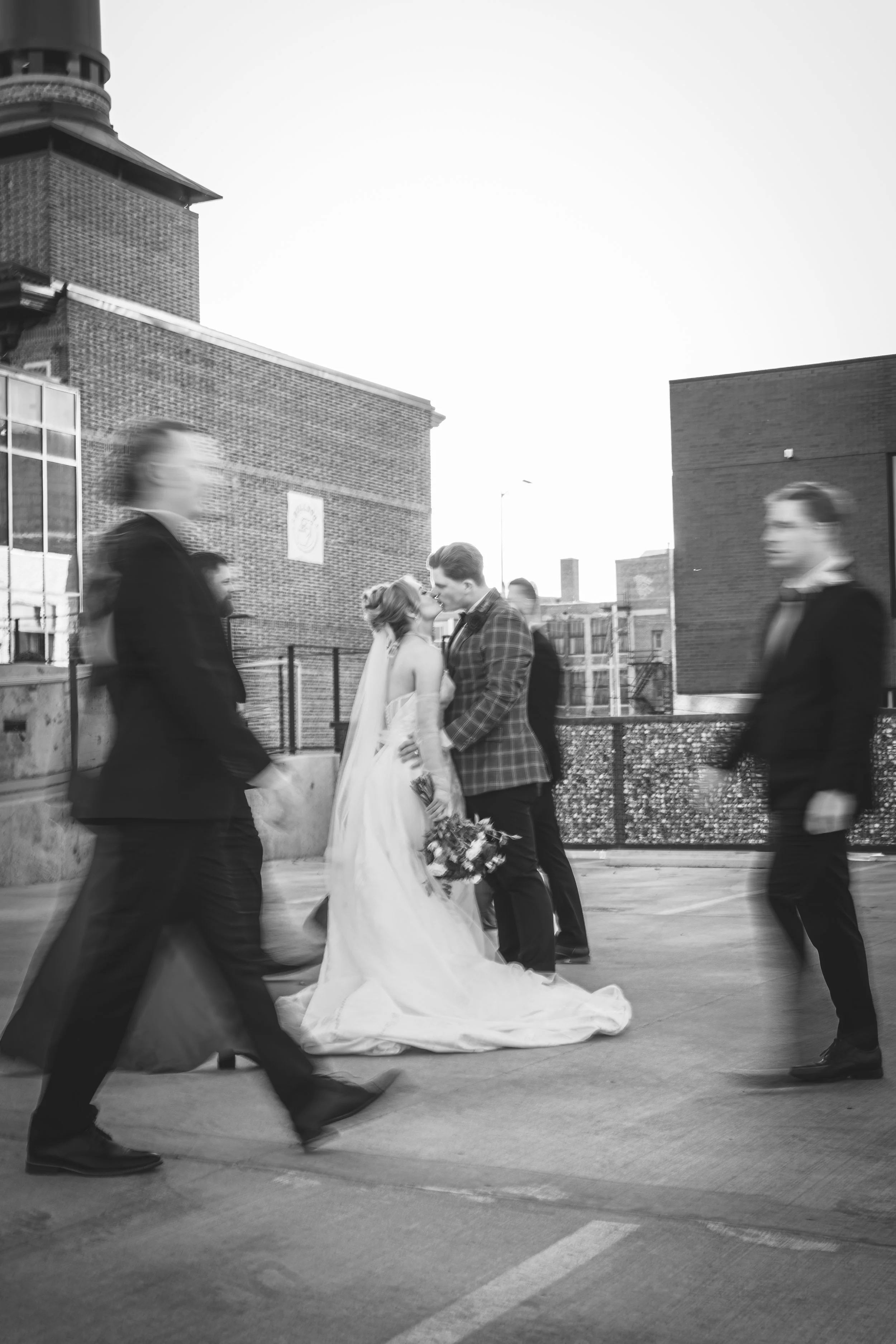 A bride and groom sharing a kiss while walking on a city street surrounded by blurred pedestrians. The bride is wearing a wedding gown with a long train, and the groom is in a checkered suit. The scene is black and white.