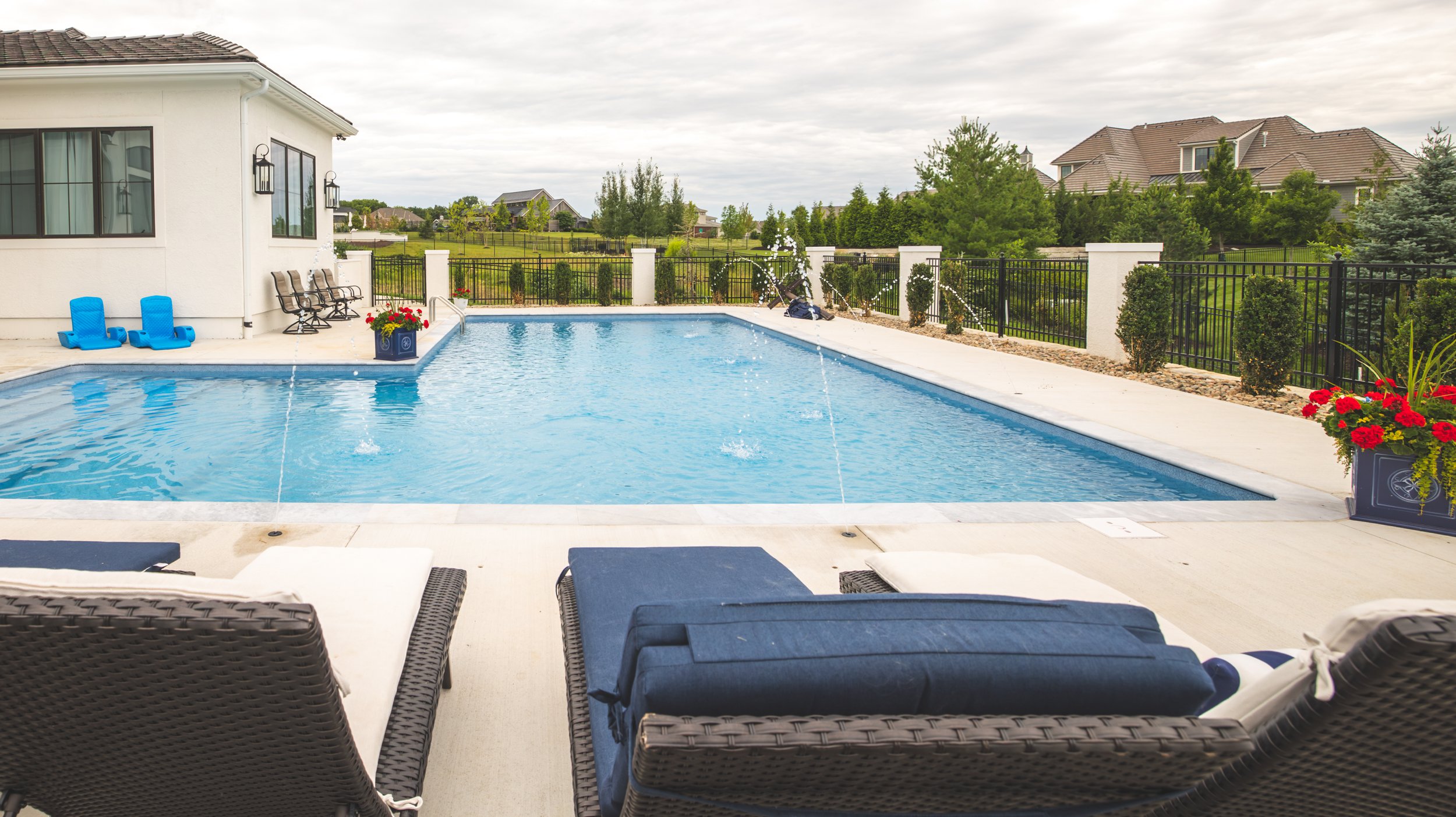 View of a backyard swimming pool with four lounge chairs and two blue floaties, surrounded by a white fence and landscaped with bushes and flowers, with neighboring houses and trees in the background.