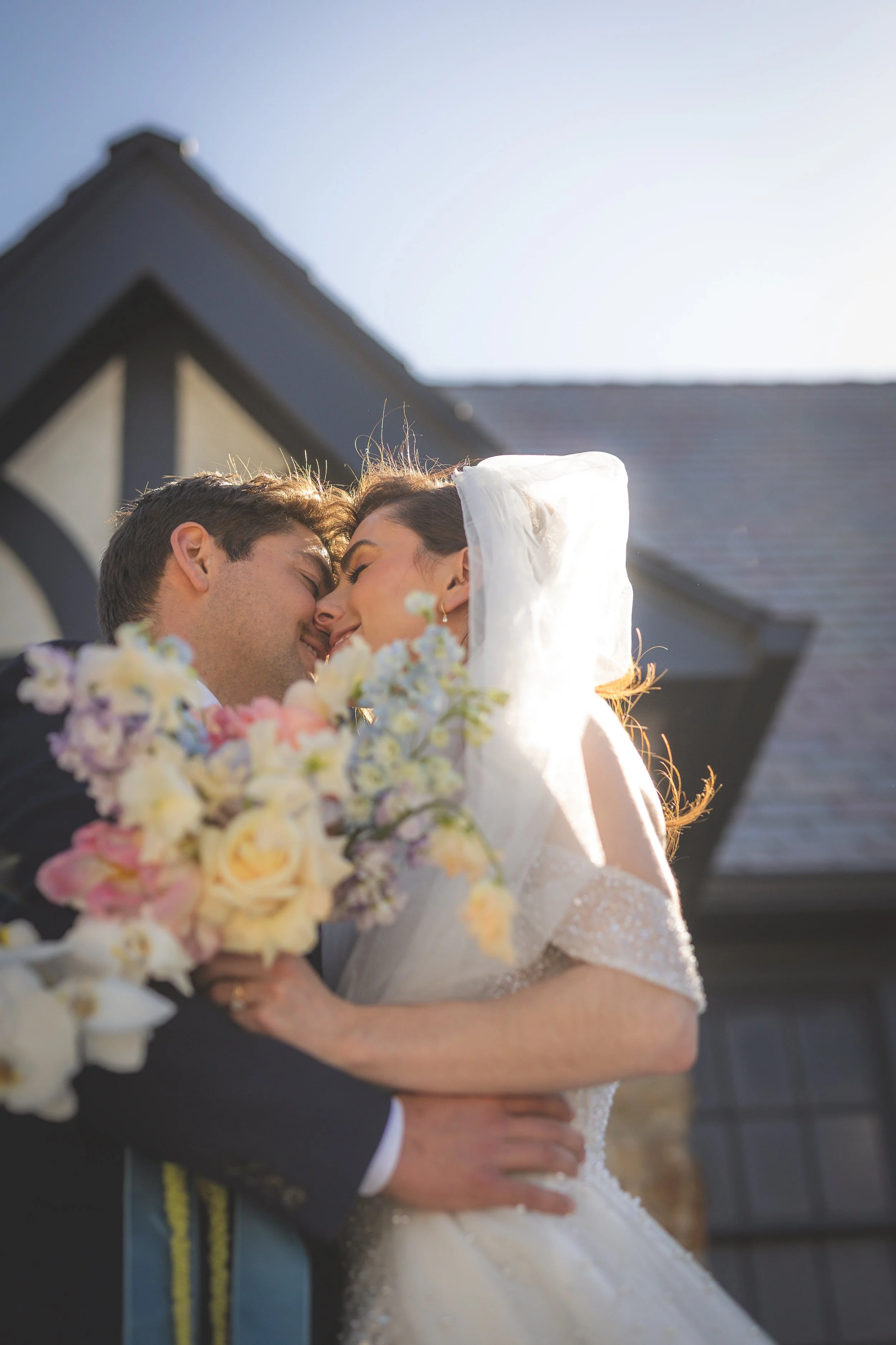 A bride and groom are embracing during their wedding, with their foreheads touching and eyes closed, standing outside near a house with a gabled roof.