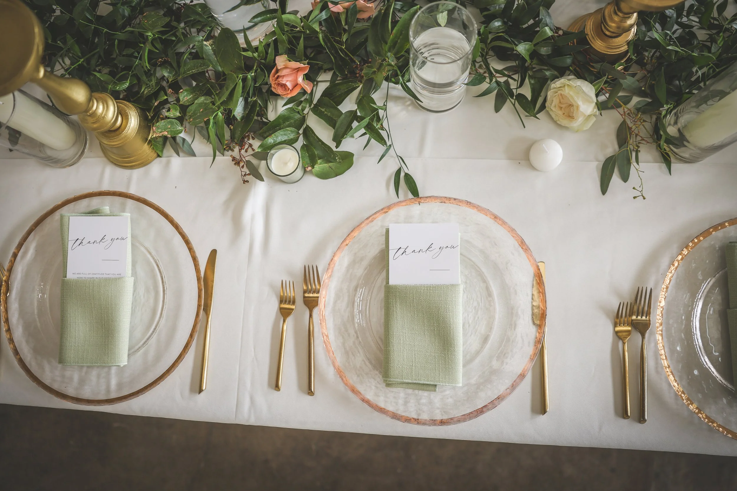 Elegant dining table with three place settings featuring glass plates with rose gold rims, light green napkins, and gold utensils. The table is decorated with greenery, pink and white roses, candles, and gold candle holders.