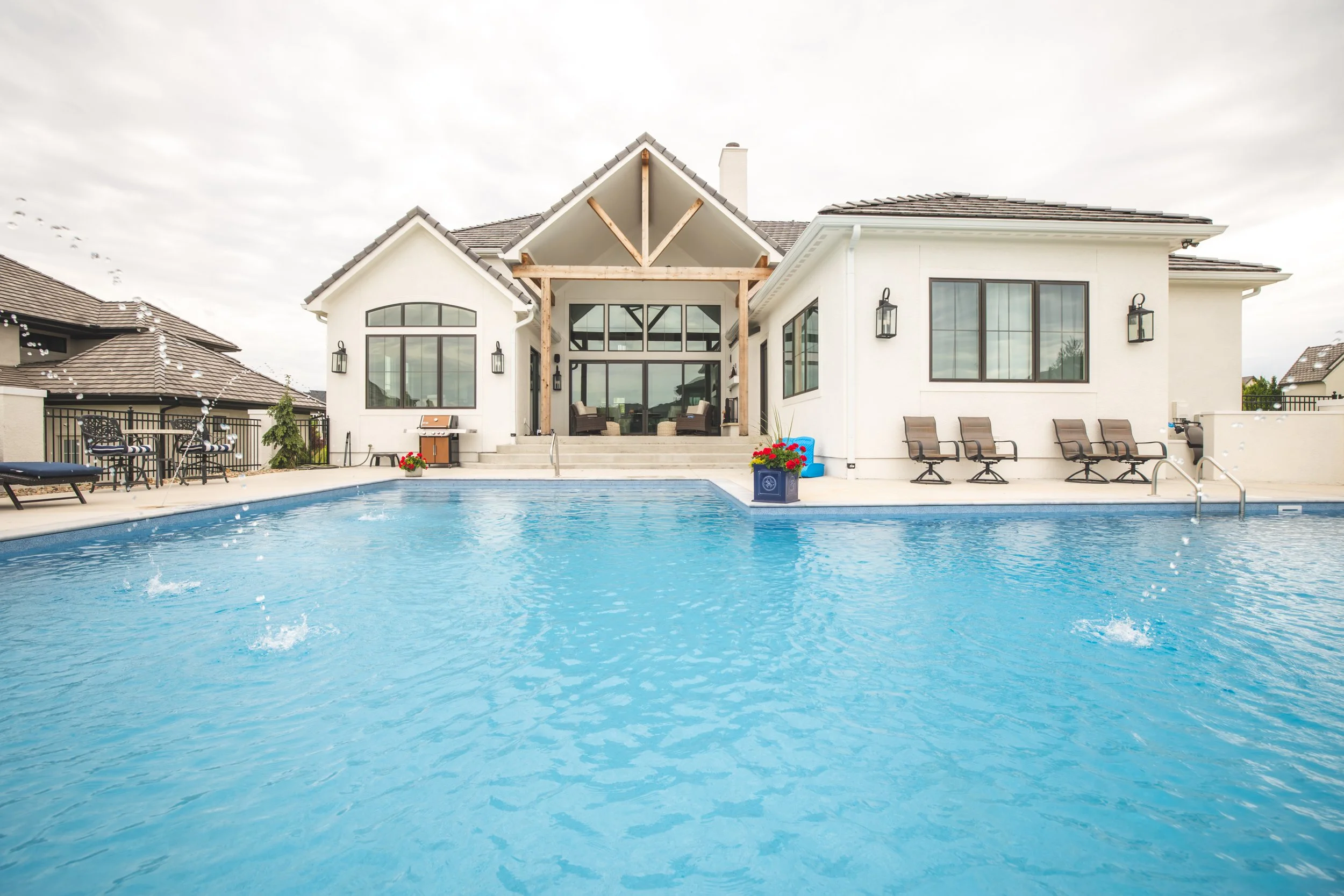 A backyard with a swimming pool in front of a modern white house with large windows and outdoor seating, under cloudy skies.