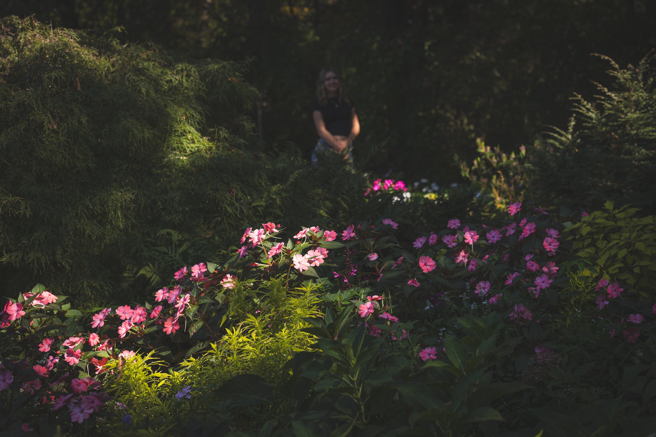 A woman standing in a lush garden with pink, purple, and white flowers, surrounded by green foliage and trees, with sunlight filtering through.