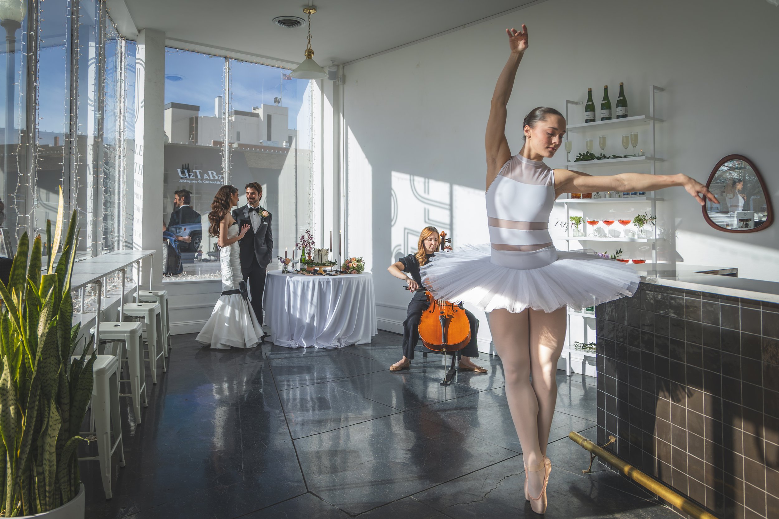 A ballet dancer in a white tutu and leotard practicing at a dance studio or restaurant. In the background, a couple dressed in wedding attire is talking near a table with flowers, and a woman is playing a cello. The studio has large windows, white wa