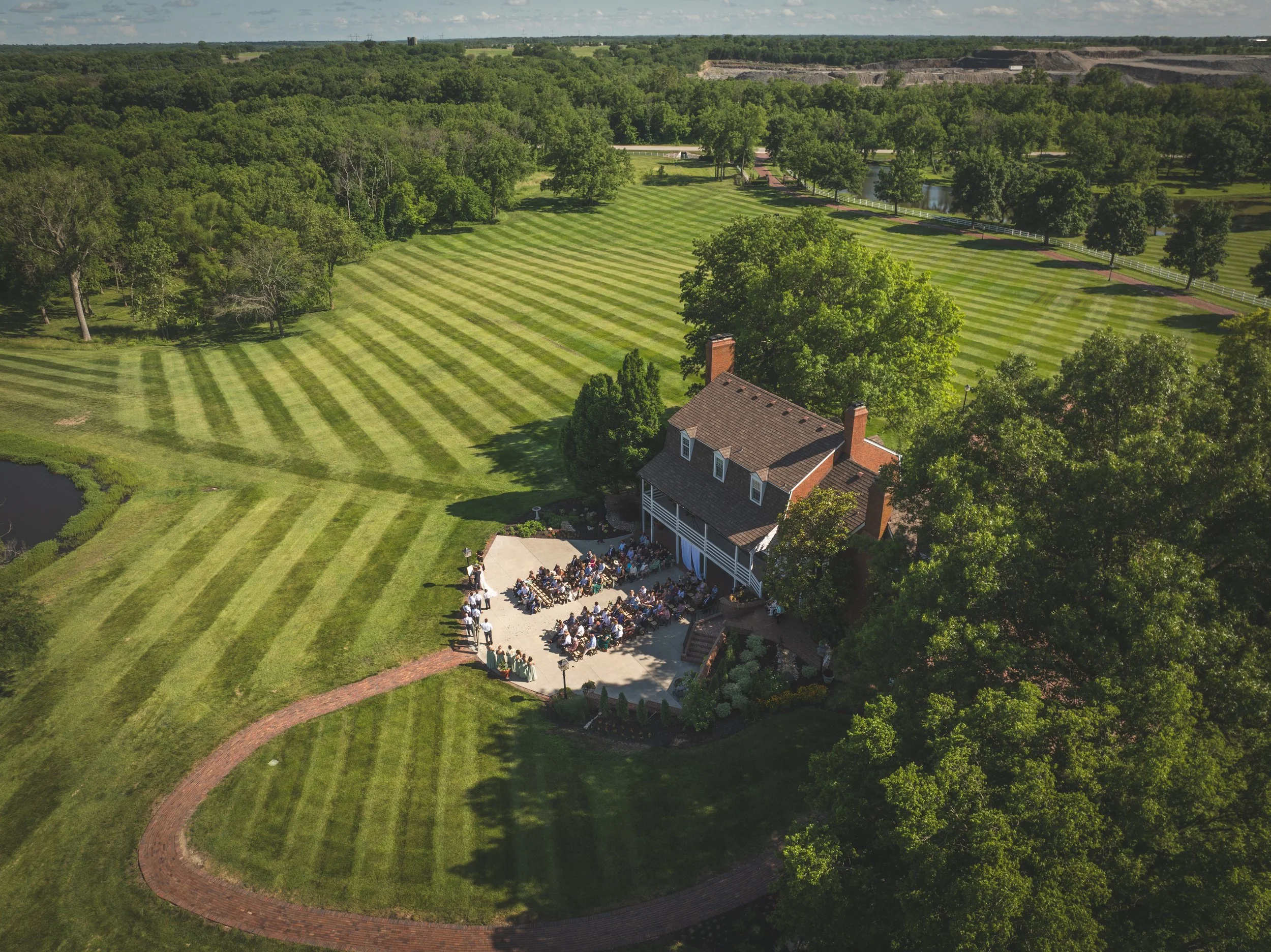 Aerial view of a large outdoor gathering with many people seated on a patio in front of a brick house, surrounded by green grass, trees, and a vast field with diagonal striped mowing lines.