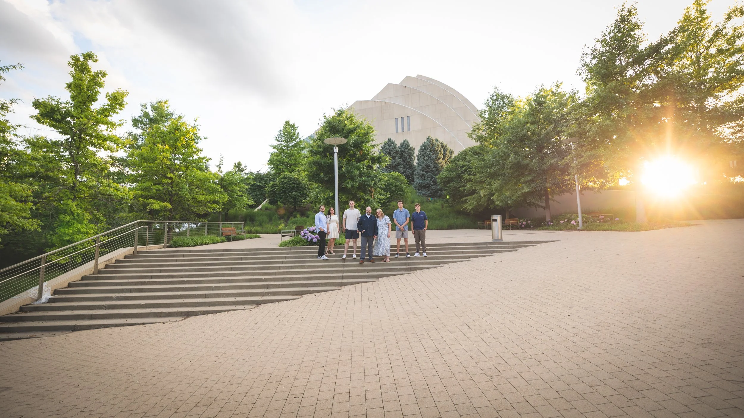 A group of seven people standing on steps outdoors with trees and a modern building in background, illuminated by bright sunlight.