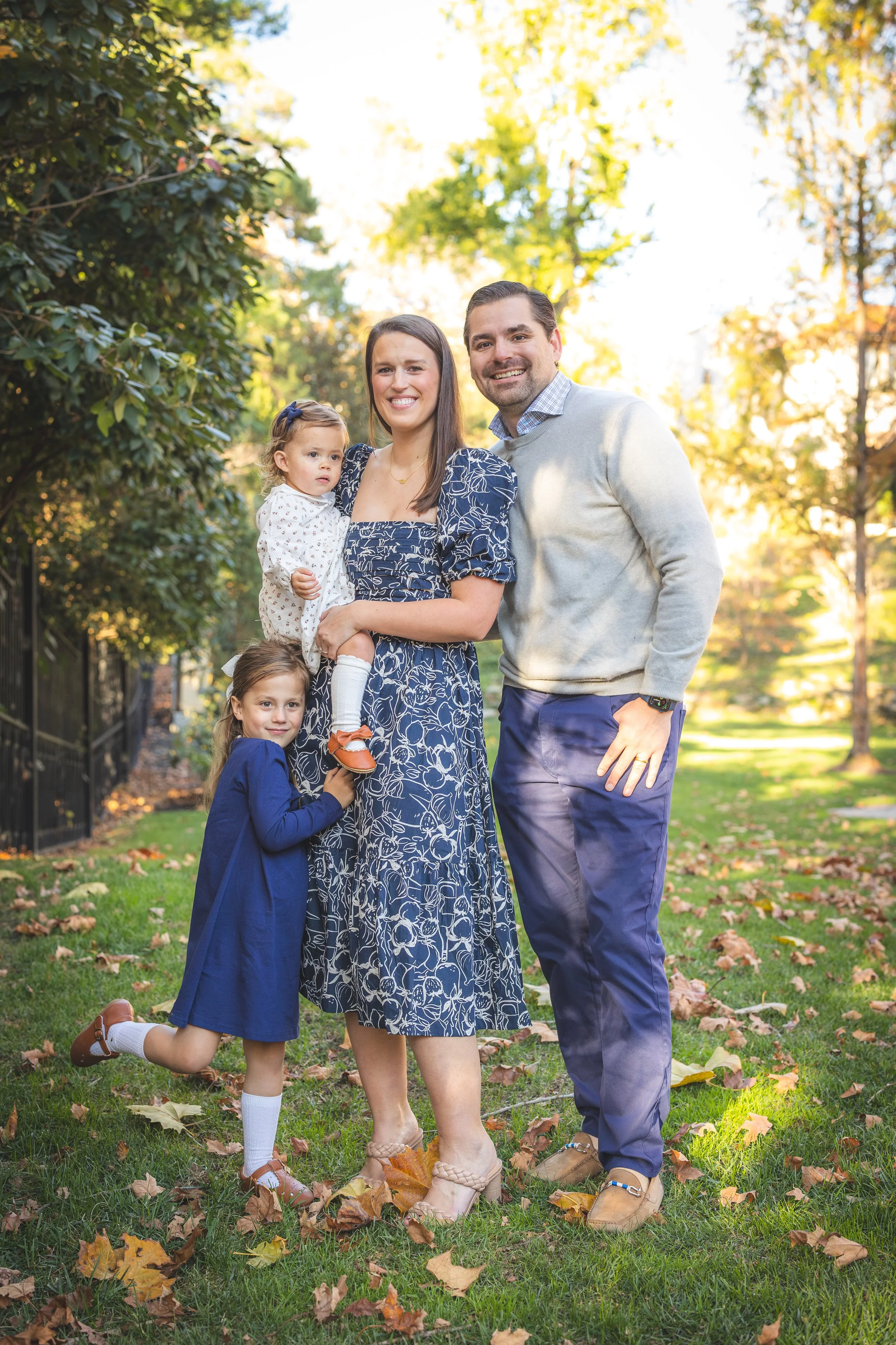 A family of four, including two young girls and two adults, standing together outdoors on a sunny autumn day with fallen leaves on the grass.