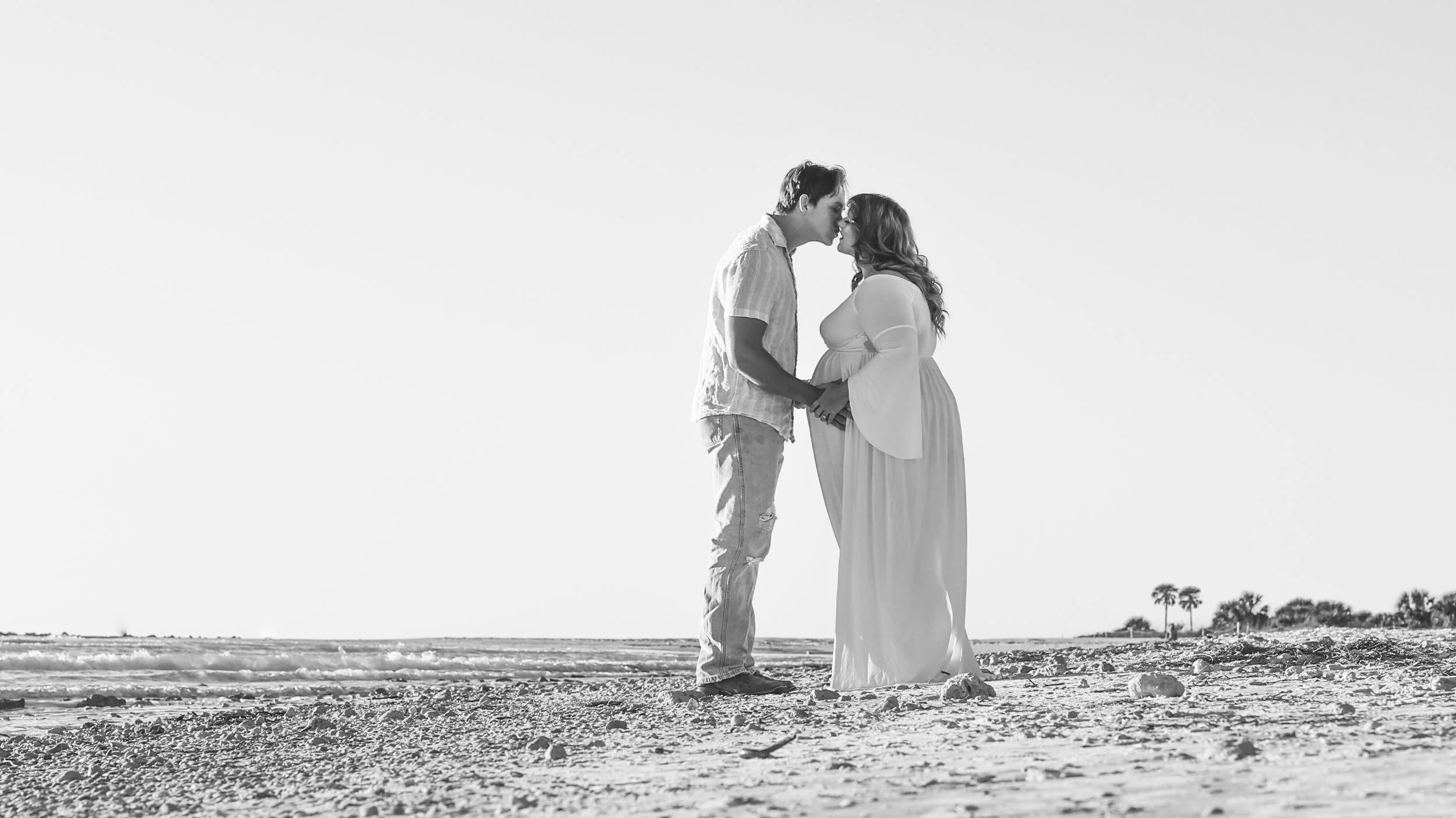 A black and white photo of a couple holding hands and touching foreheads on a beach, with the ocean and palm trees in the background.