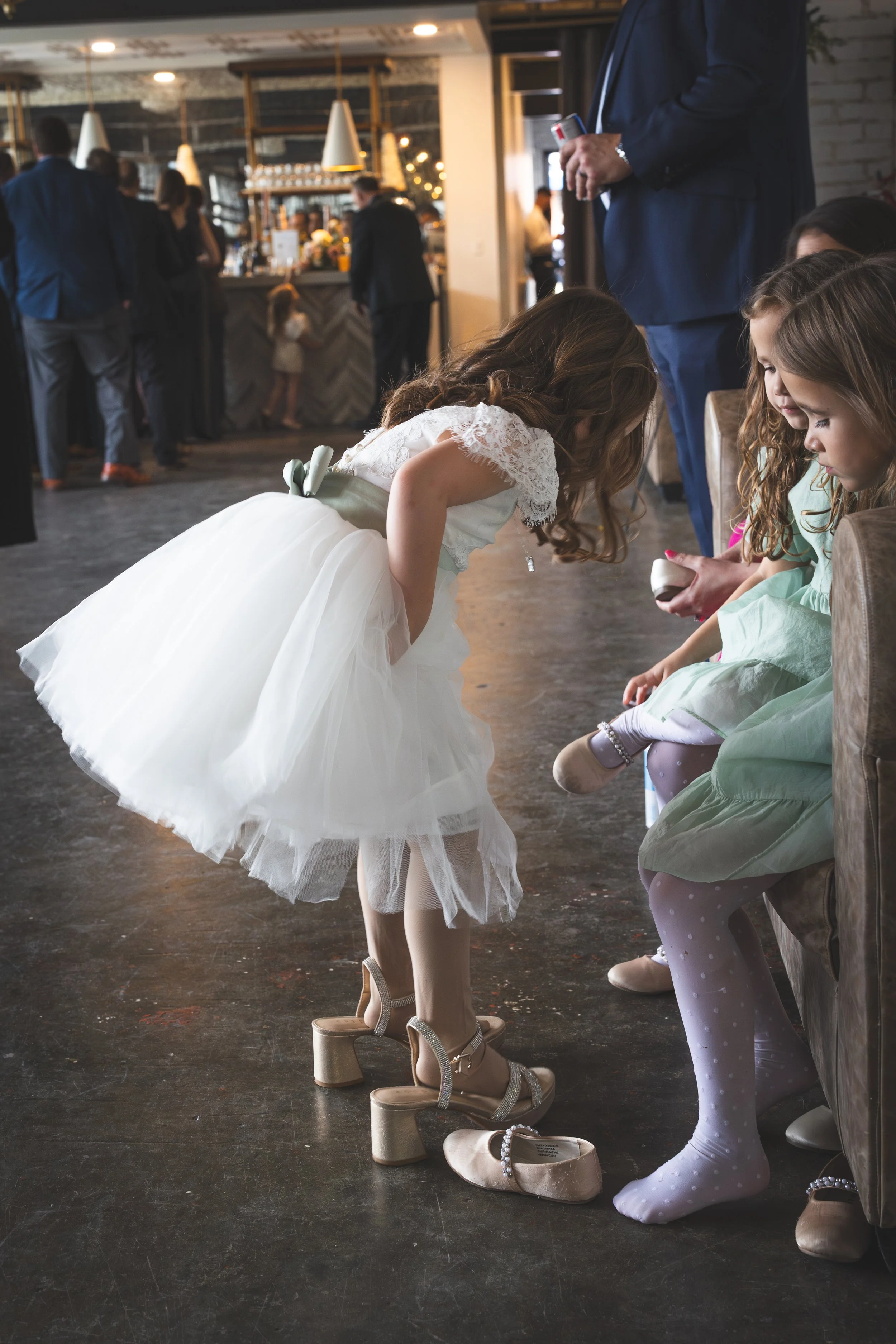 Children sitting on a bench and looking at shoes on the floor at a wedding reception, with adults standing in the background and a bar area in a rustic venue.