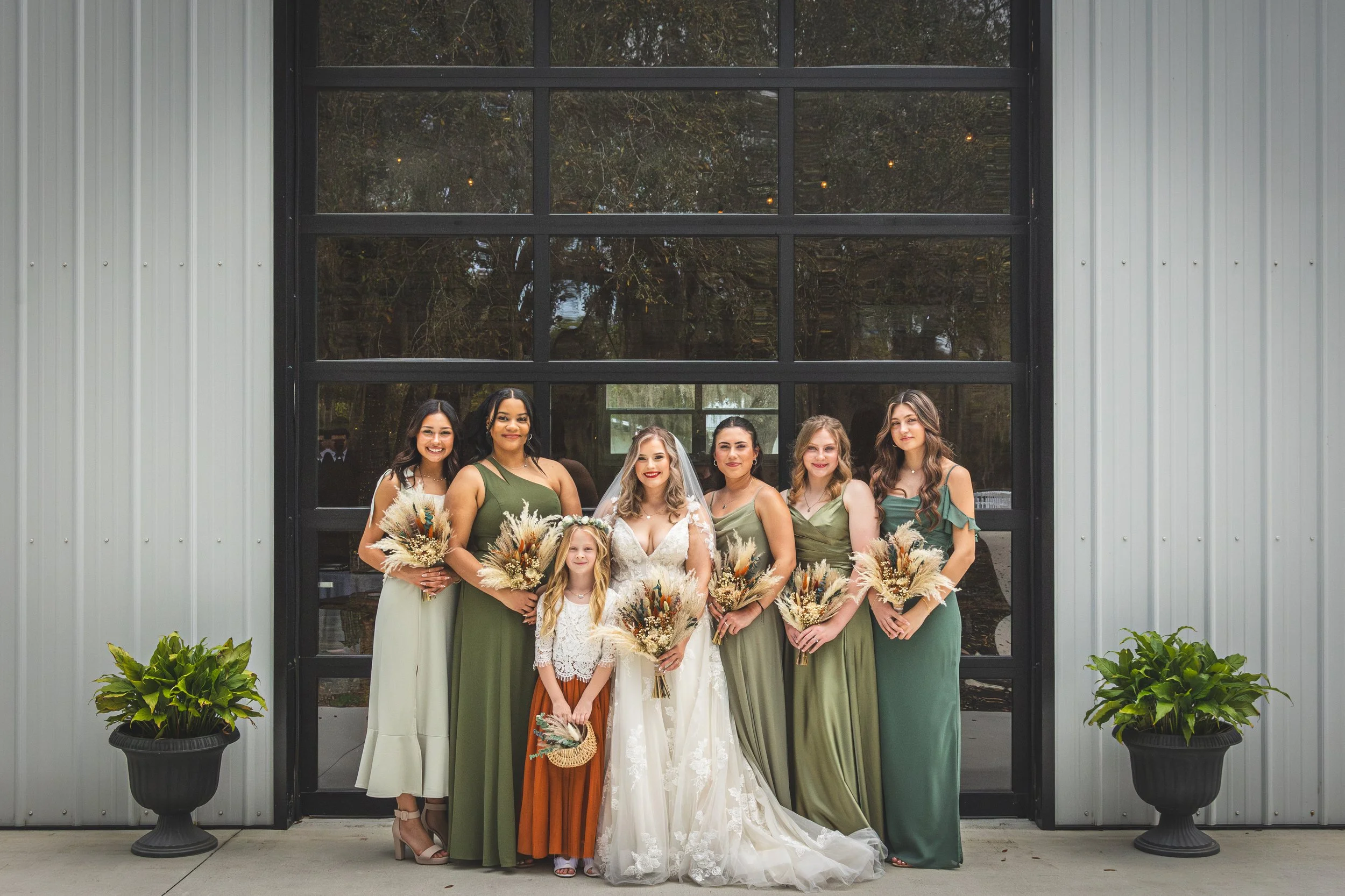 A group of women and young girl dressed in formal attire, standing in front of a large glass window and potted plants, during a wedding celebration.