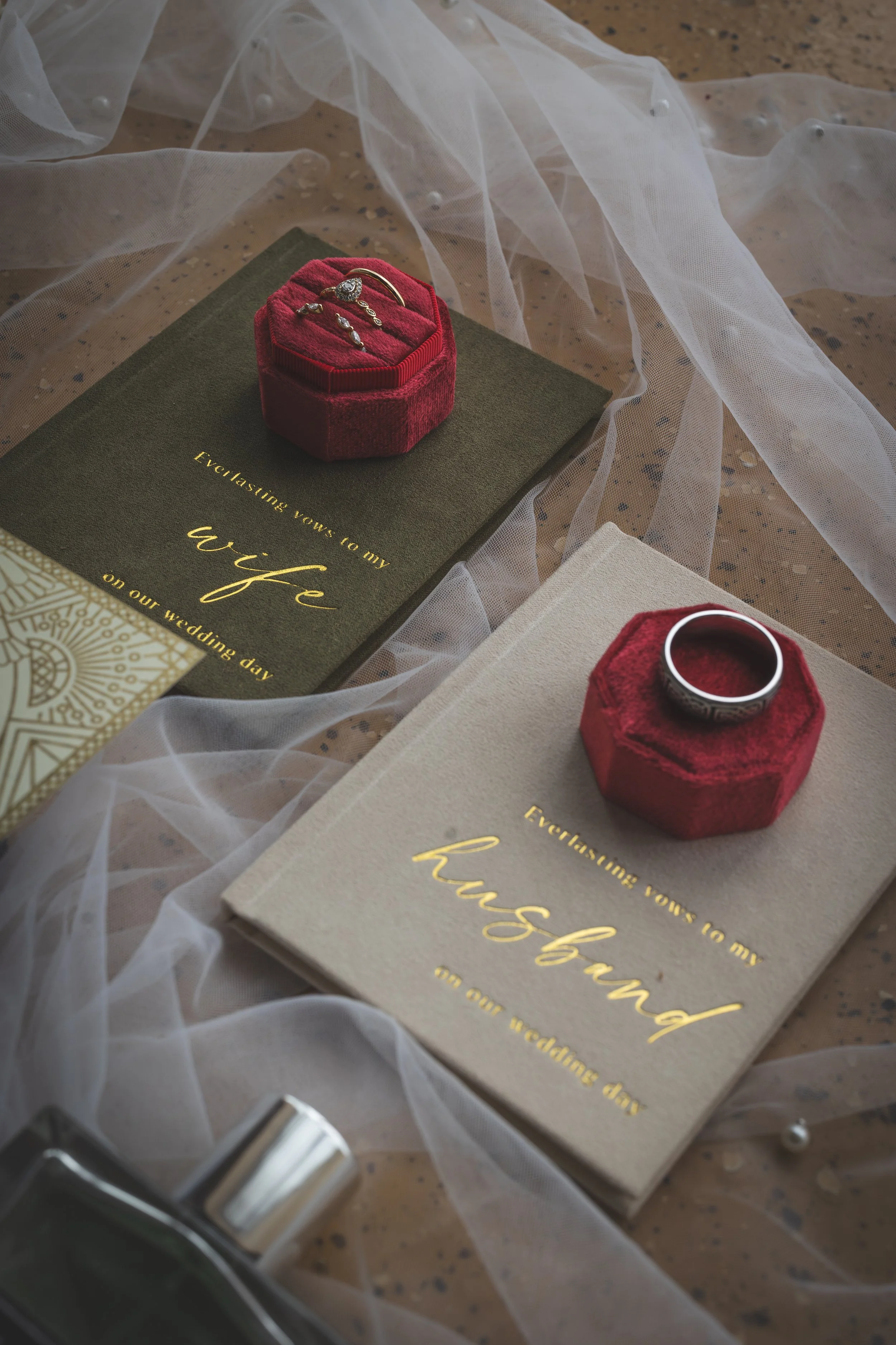 Pink velvet jewelry box with rings and earrings on top of a wedding invitation card with gold lettering, surrounded by white tulle fabric and pearls, on a wooden surface.