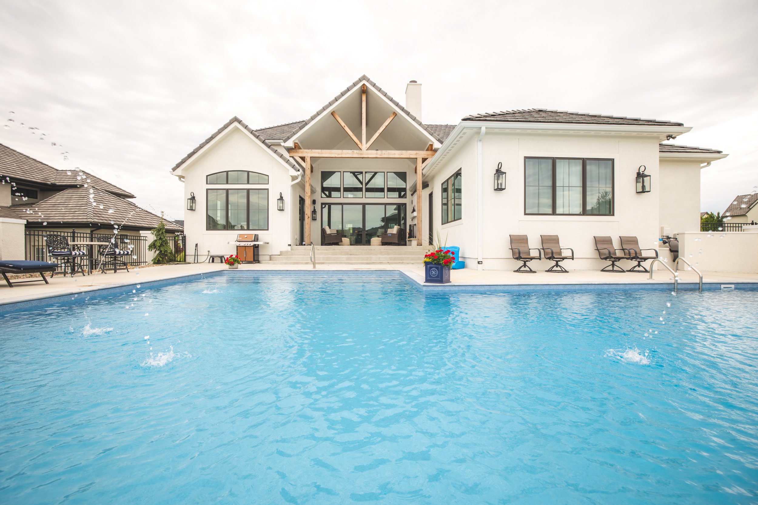 A large white house with a swimming pool in the backyard, patio chairs, and patio area with steps leading to the house, under a cloudy sky.