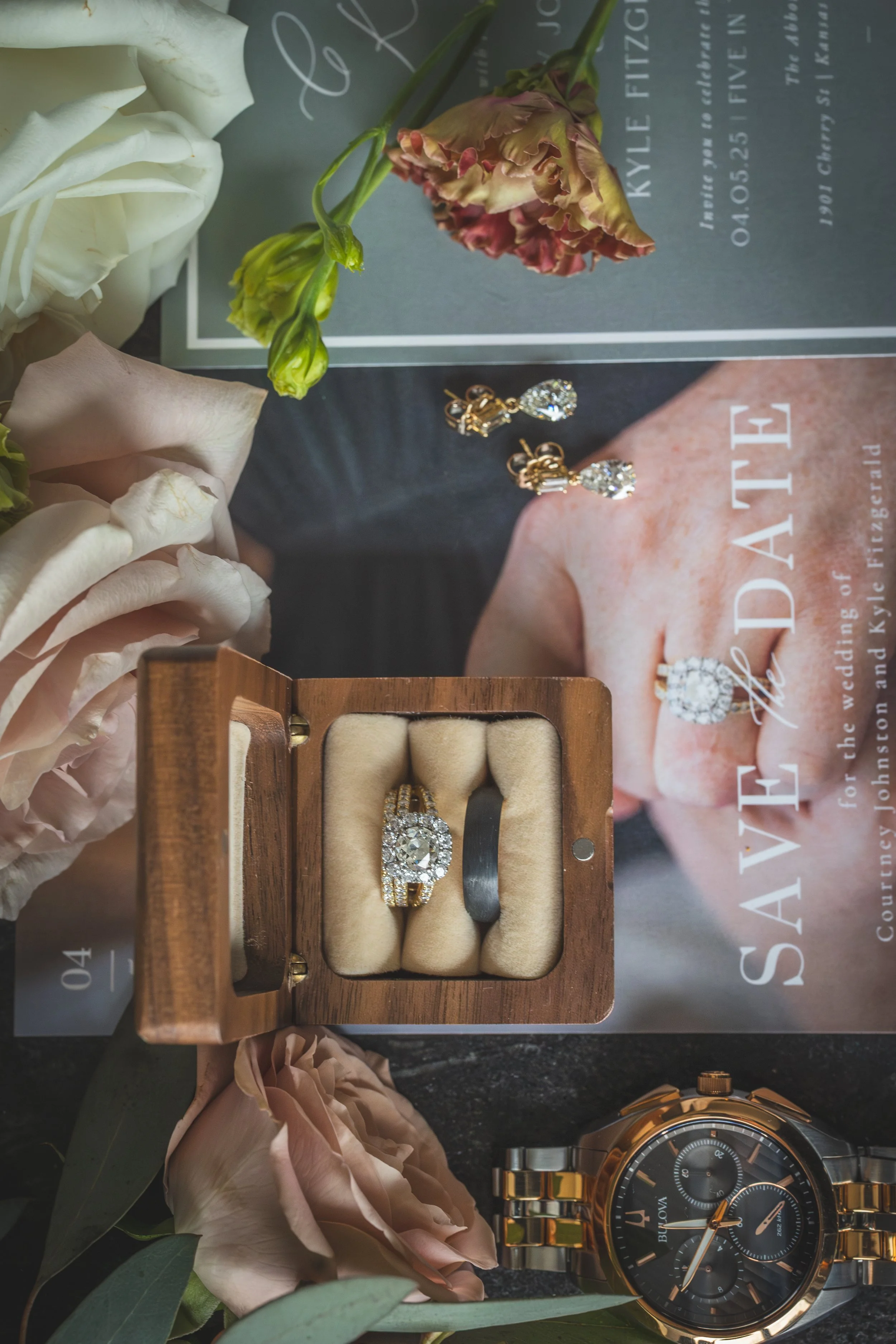 A jewelry display featuring a pair of diamond earrings, a diamond ring, and a gold watch surrounded by pink and white flowers, with a promotional magazine in the background.