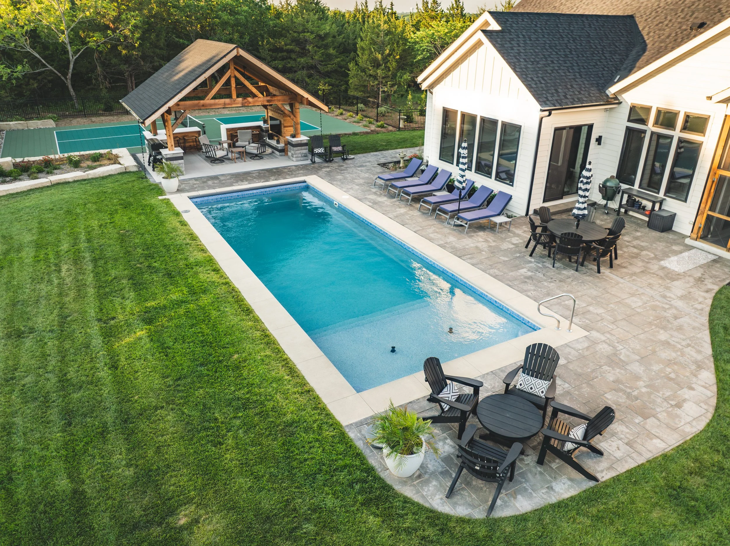A backyard with a swimming pool, a seating area under a wooden pavilion, a patio with lounge chairs, a dining table with chairs, and a house with large windows, set against a backdrop of trees.