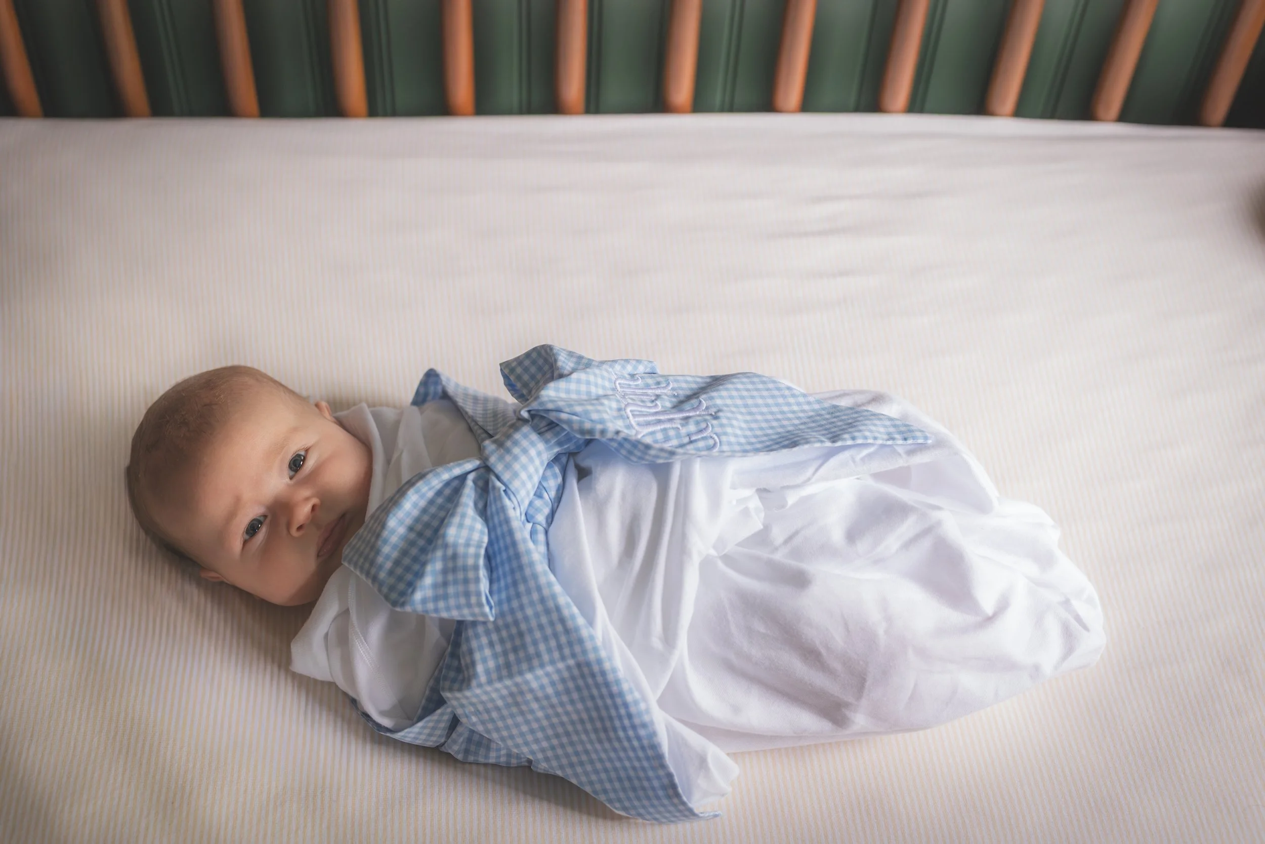 A baby lying on a bed with a striped sheet, wearing a white and blue outfit, looking at the camera.