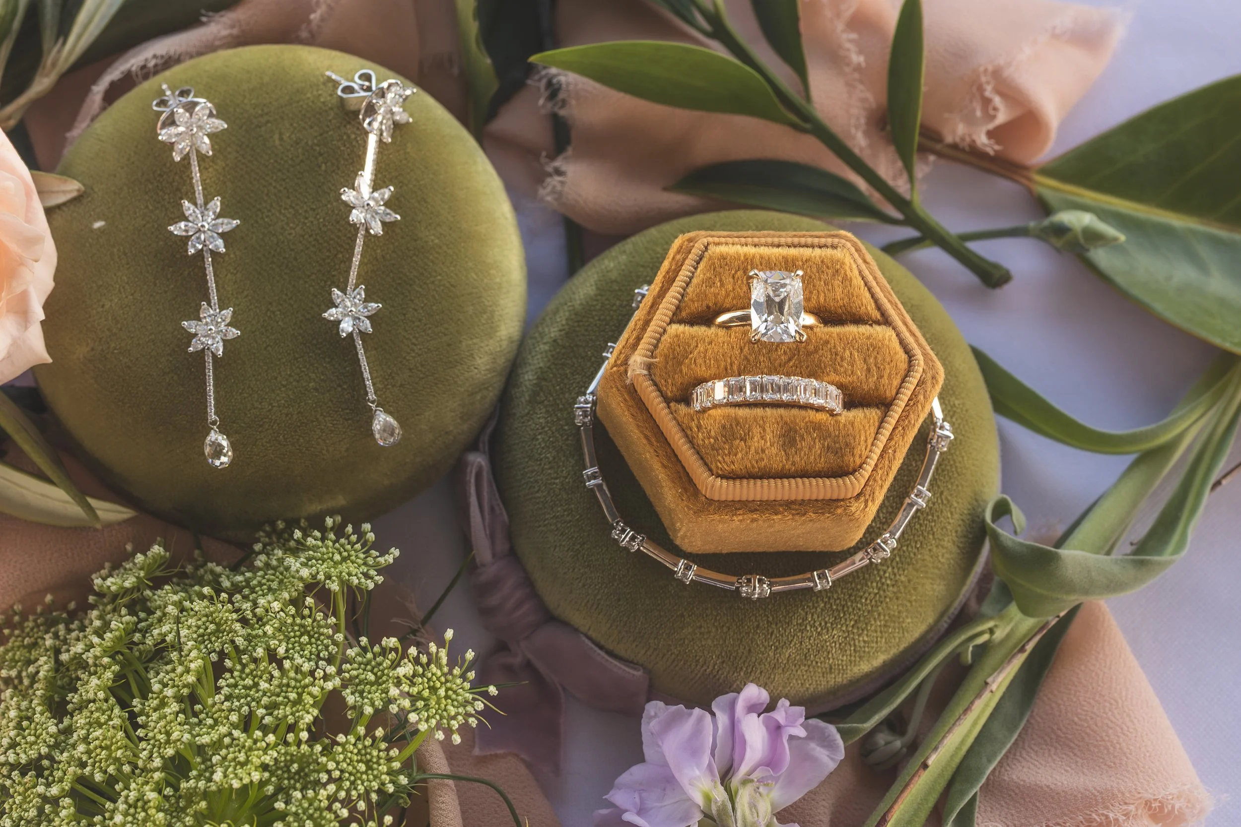 Jewelry display featuring a diamond ring in a velvet box and a pair of diamond earrings with floral design, surrounded by flowers and greenery.