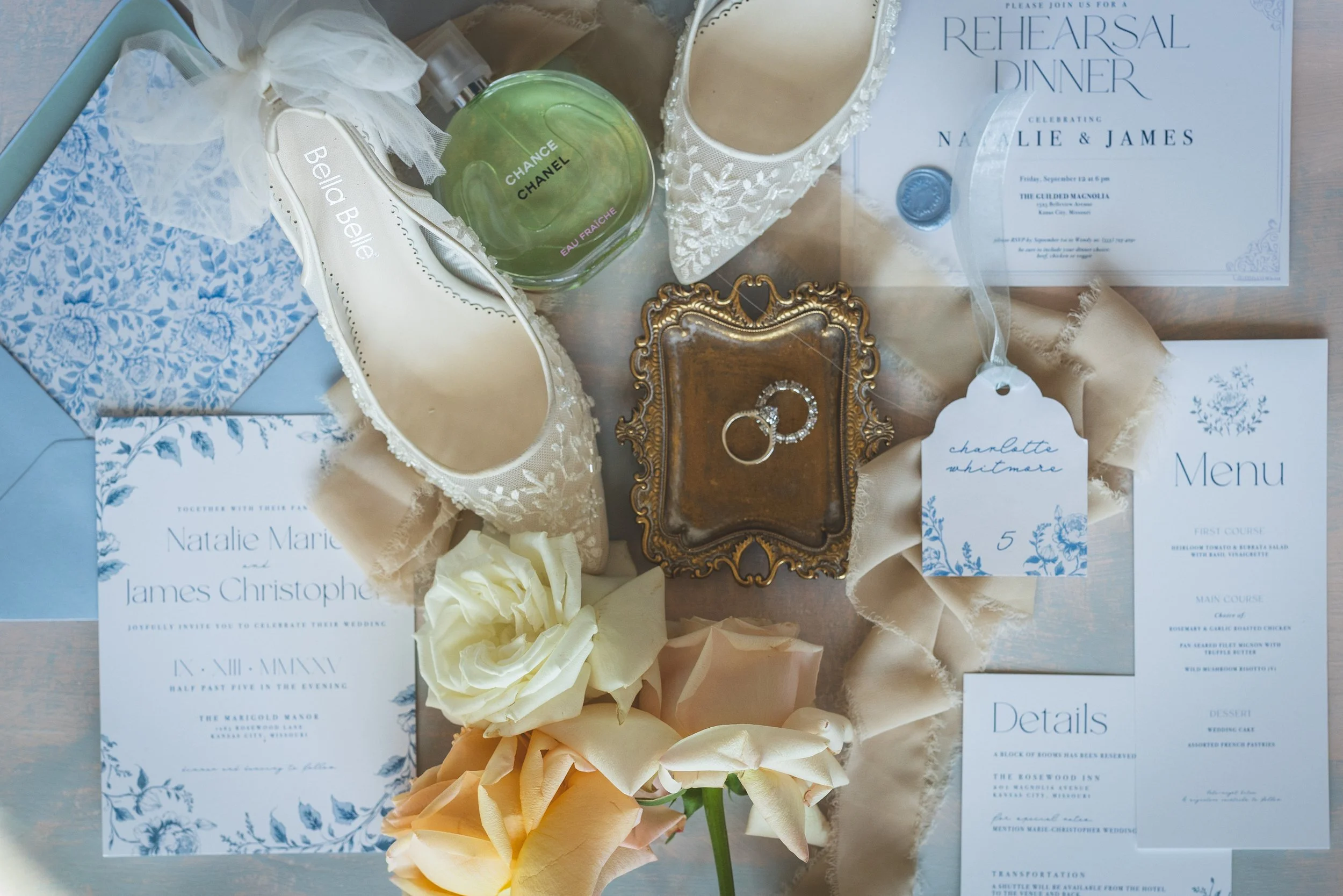 A collection of wedding items including a pair of lace wedding shoes, a bottle of Chanel Eau Fraiche perfume, a small ornate picture frame, a ring with diamonds, a white rose, a bouquet of roses, wedding invitations, and an itinerary for Natalie Marie and James Christopher's wedding rehearsal dinner.