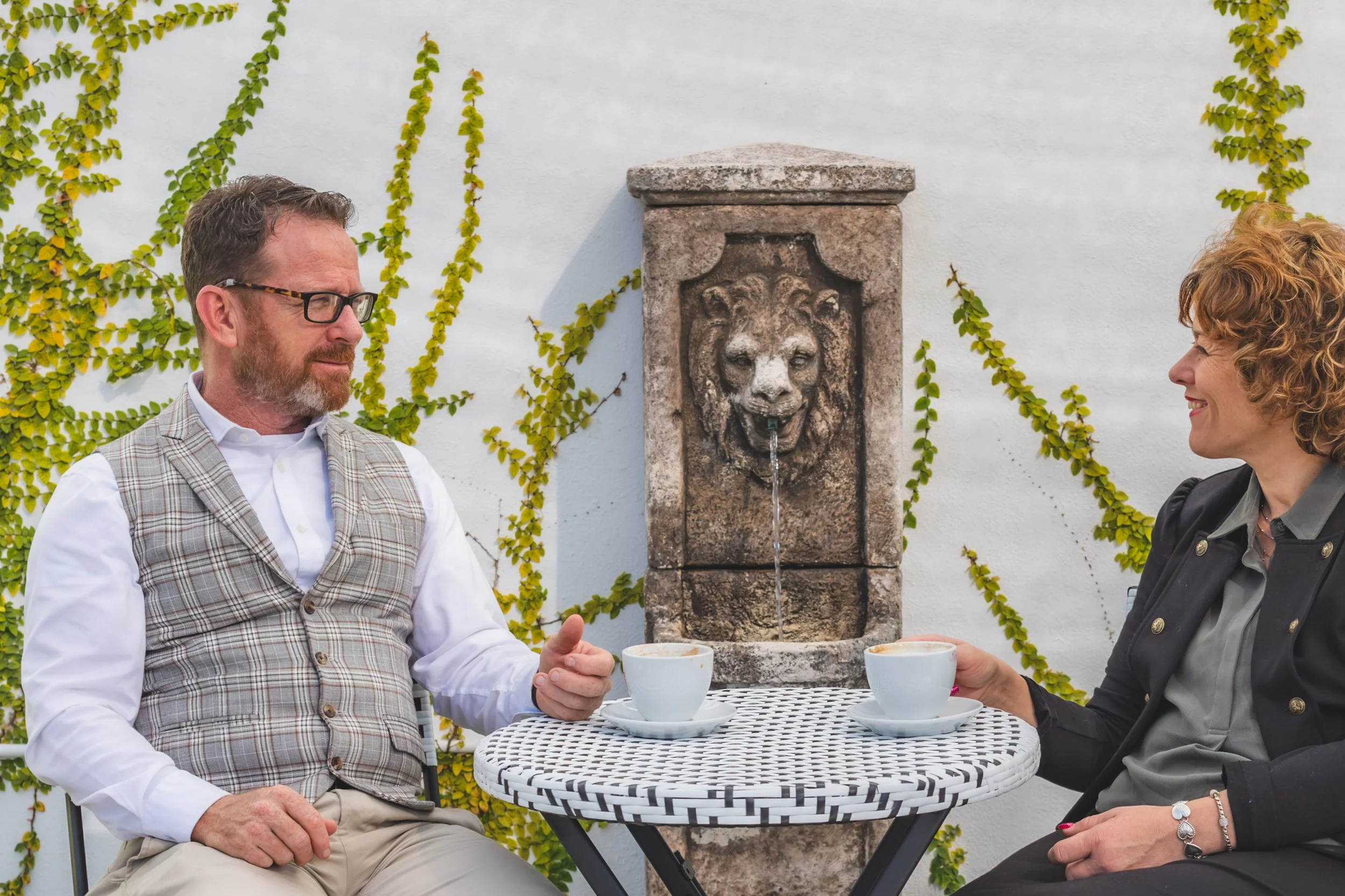 A man and woman sitting at a small round table outdoors, having coffee, with a stone fountain featuring a lion's head on the white wall behind them, surrounded by green vine plants.