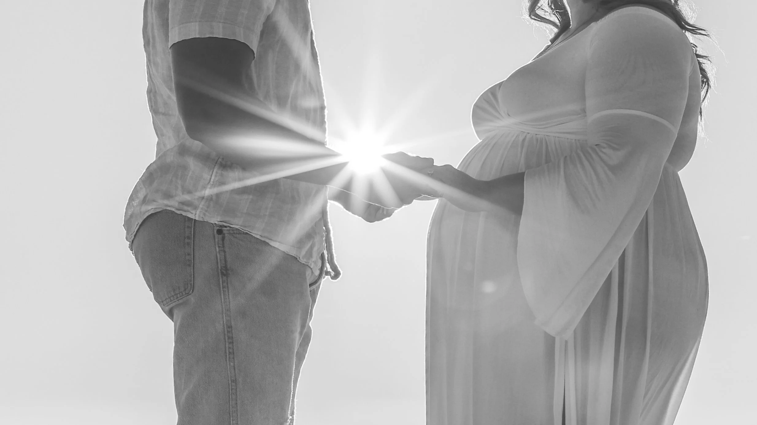 A pregnant woman and a man are holding hands, with the woman's baby bump visible, backlit by the sun creating a halo effect.
