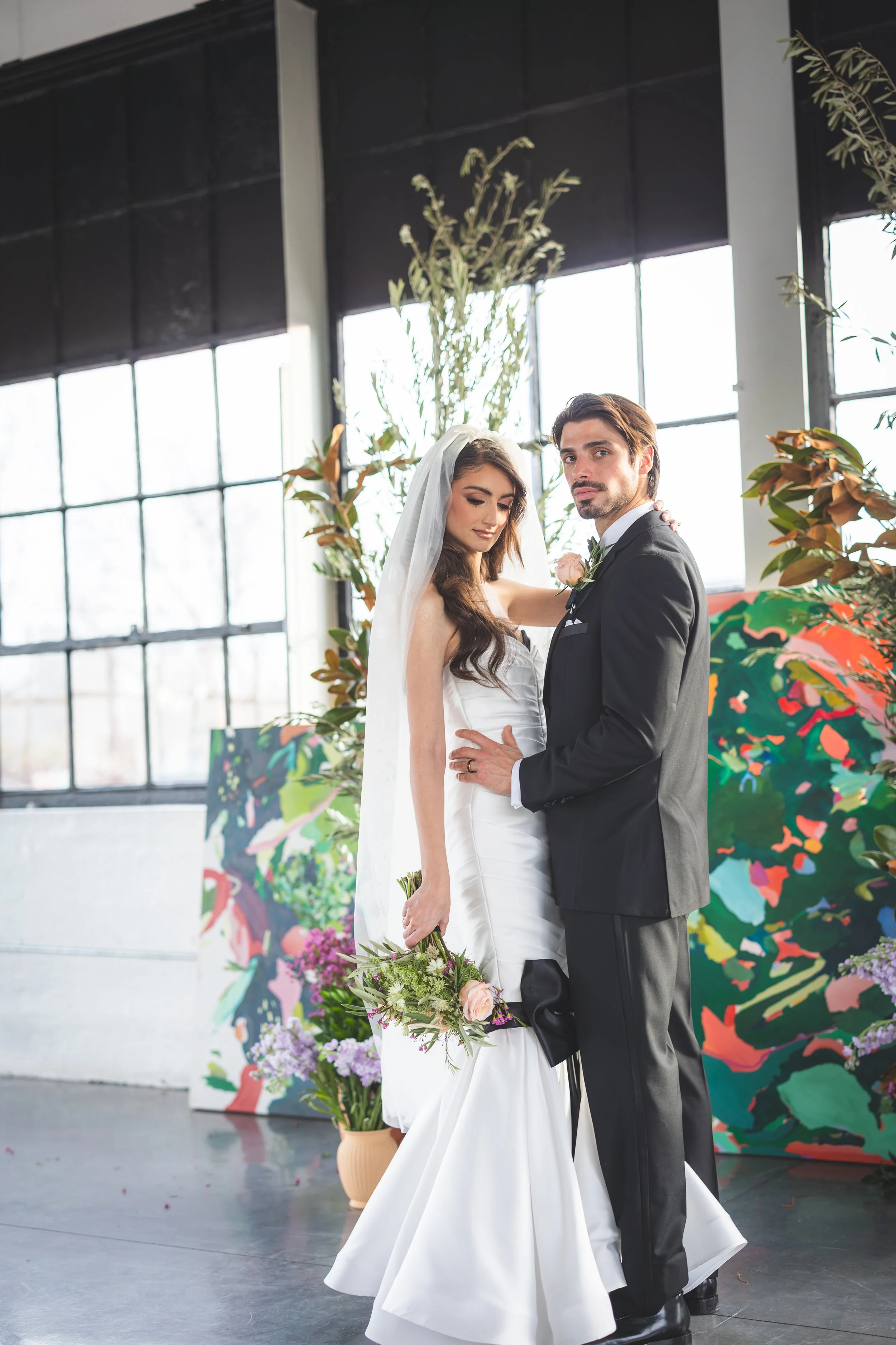 A bride and groom pose together indoors, with large windows and colorful abstract art in the background. The bride wears a white wedding gown and veil, holding a bouquet of flowers. The groom wears a black suit with a boutonniere. They stand close, with the bride's arm around the groom's neck and the groom's hand on the bride's waist.