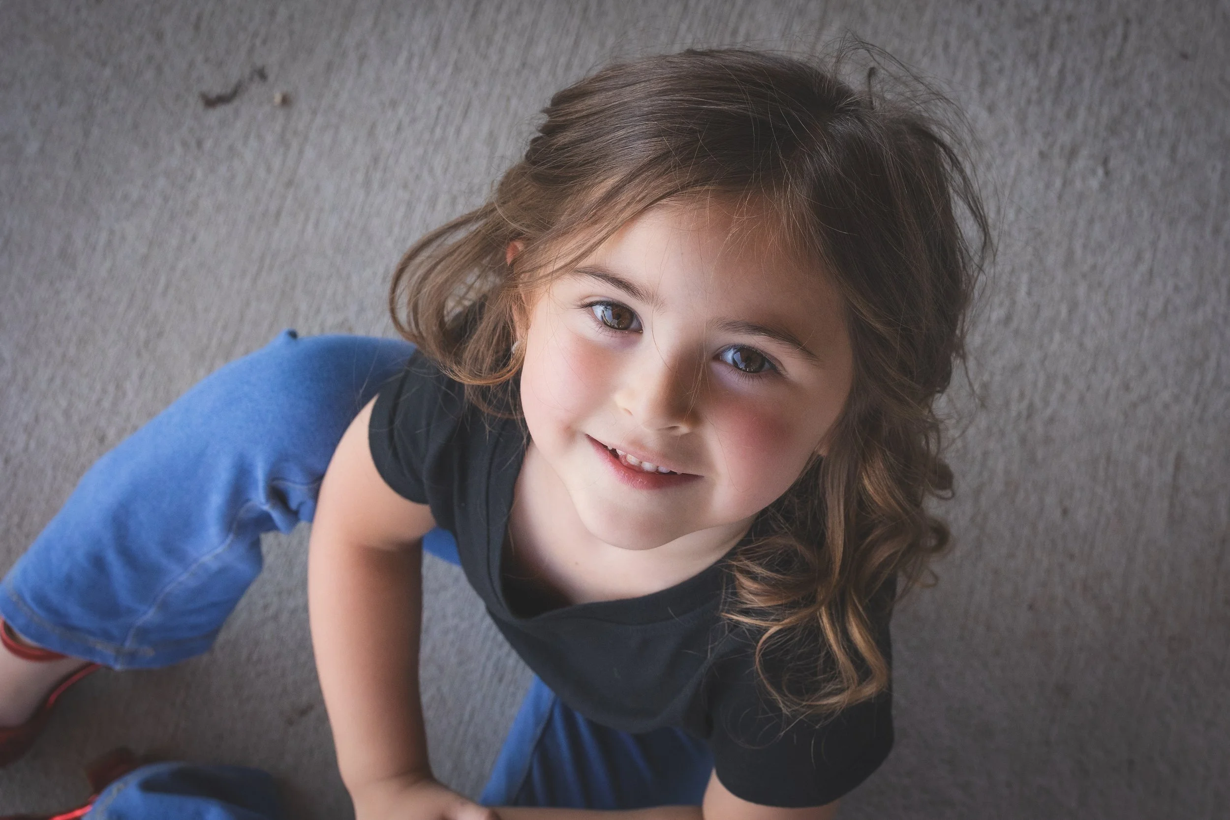 A young girl with brown, wavy hair and brown eyes, wearing a black tank top and blue jeans, sitting on a carpeted floor looking up at the camera and smiling.