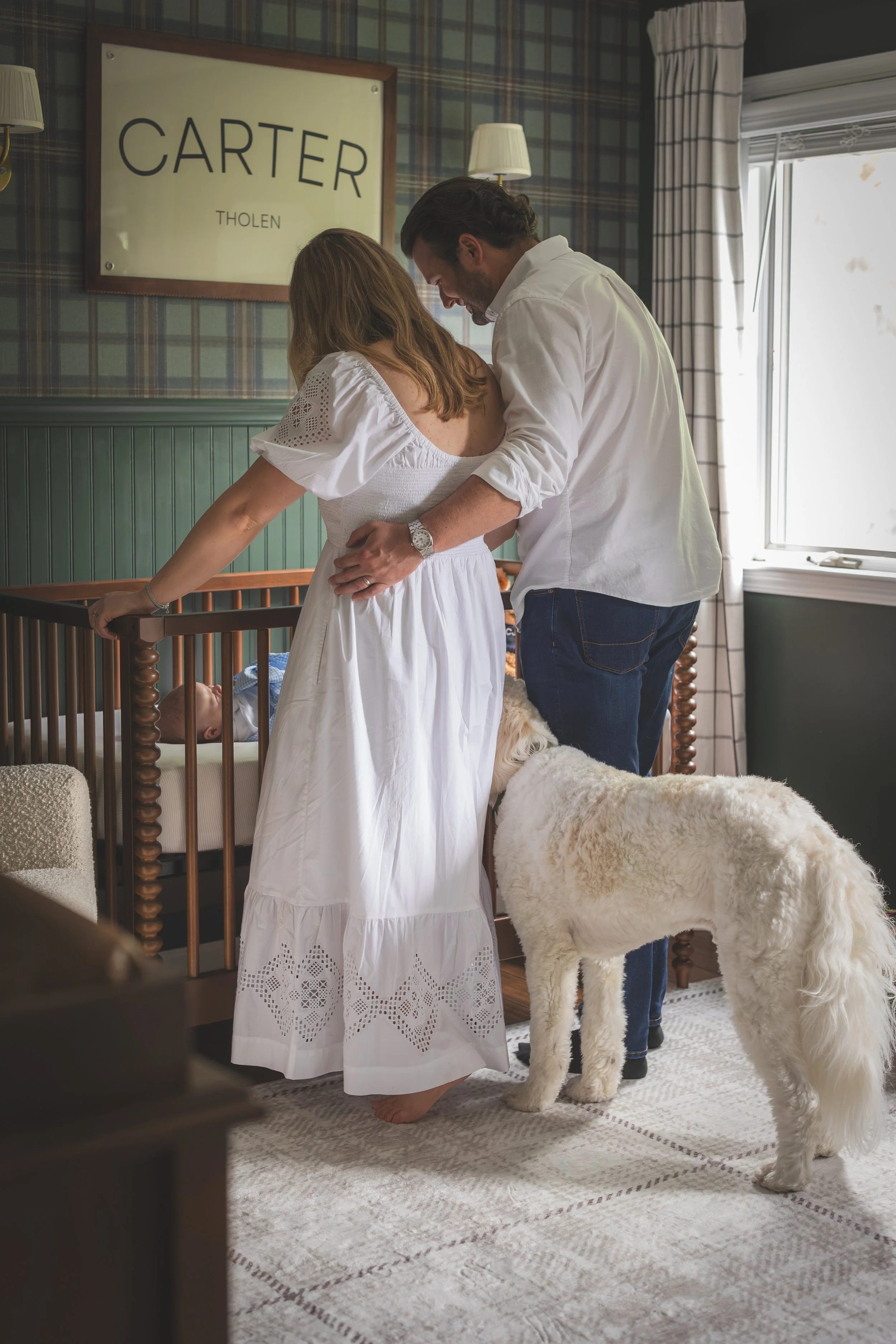 A couple stands near a crib, looking at a baby sleeping inside, in a room with plaid wallpaper, a framed sign with the name Carter Tholen, and a large window with curtains. A large dog stands nearby.
