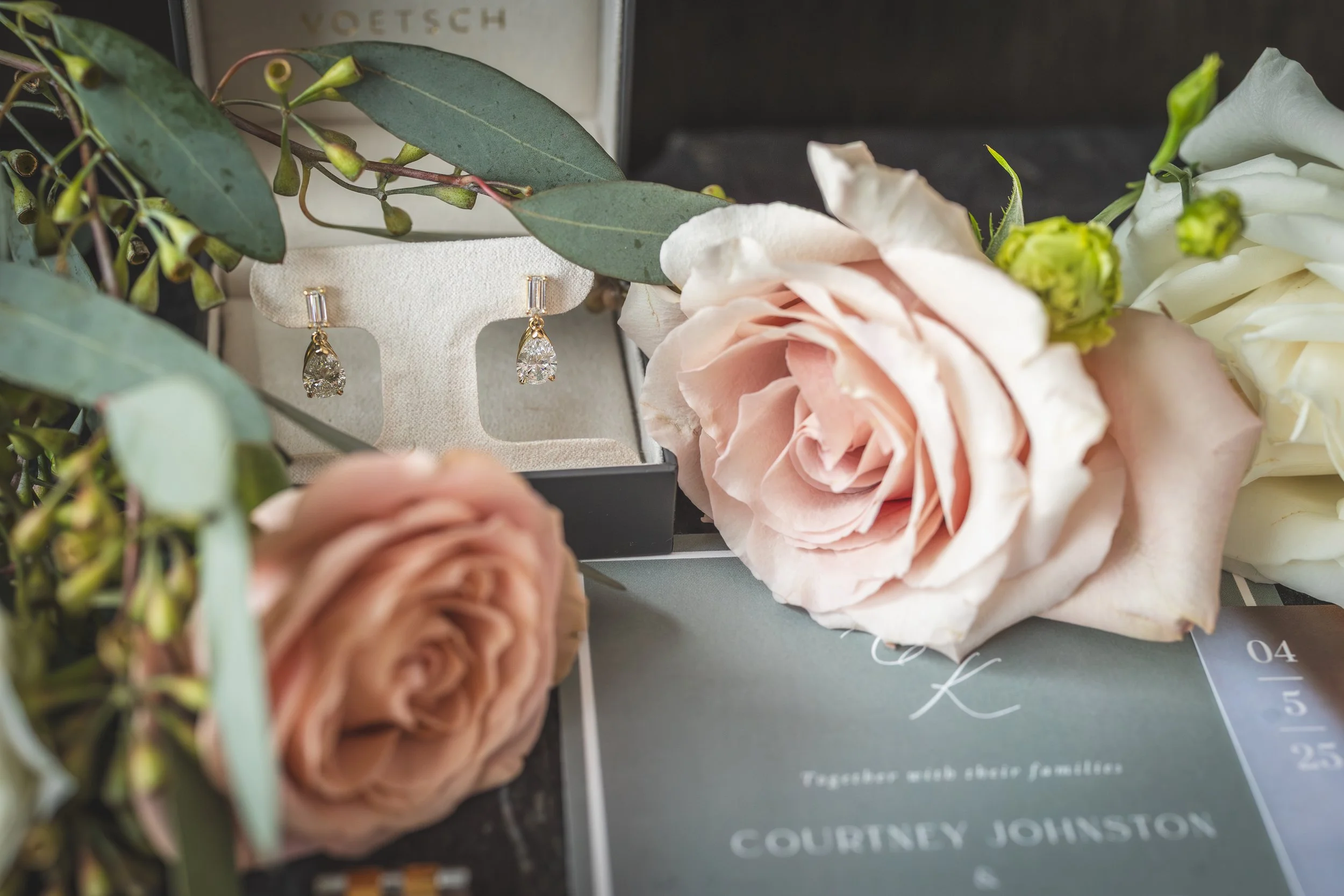 A pair of diamond earrings displayed on a jewelry stand surrounded by pink and white roses and green foliage, with a wedding invitation in the background.