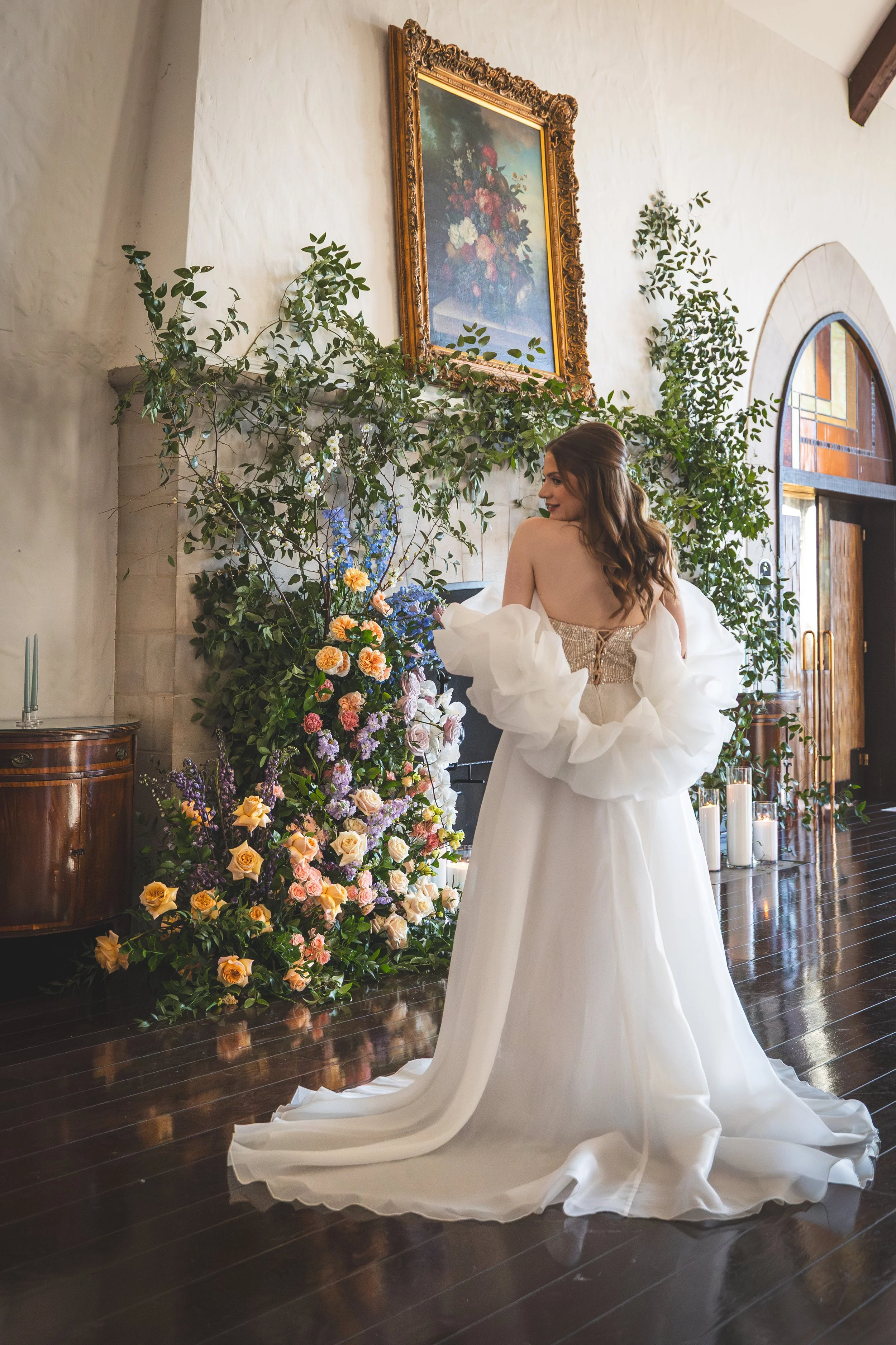 A bride in a strapless wedding gown with puffy white sleeves stands indoors in front of a decorative floral arrangement and greenery, with a framed painting above.