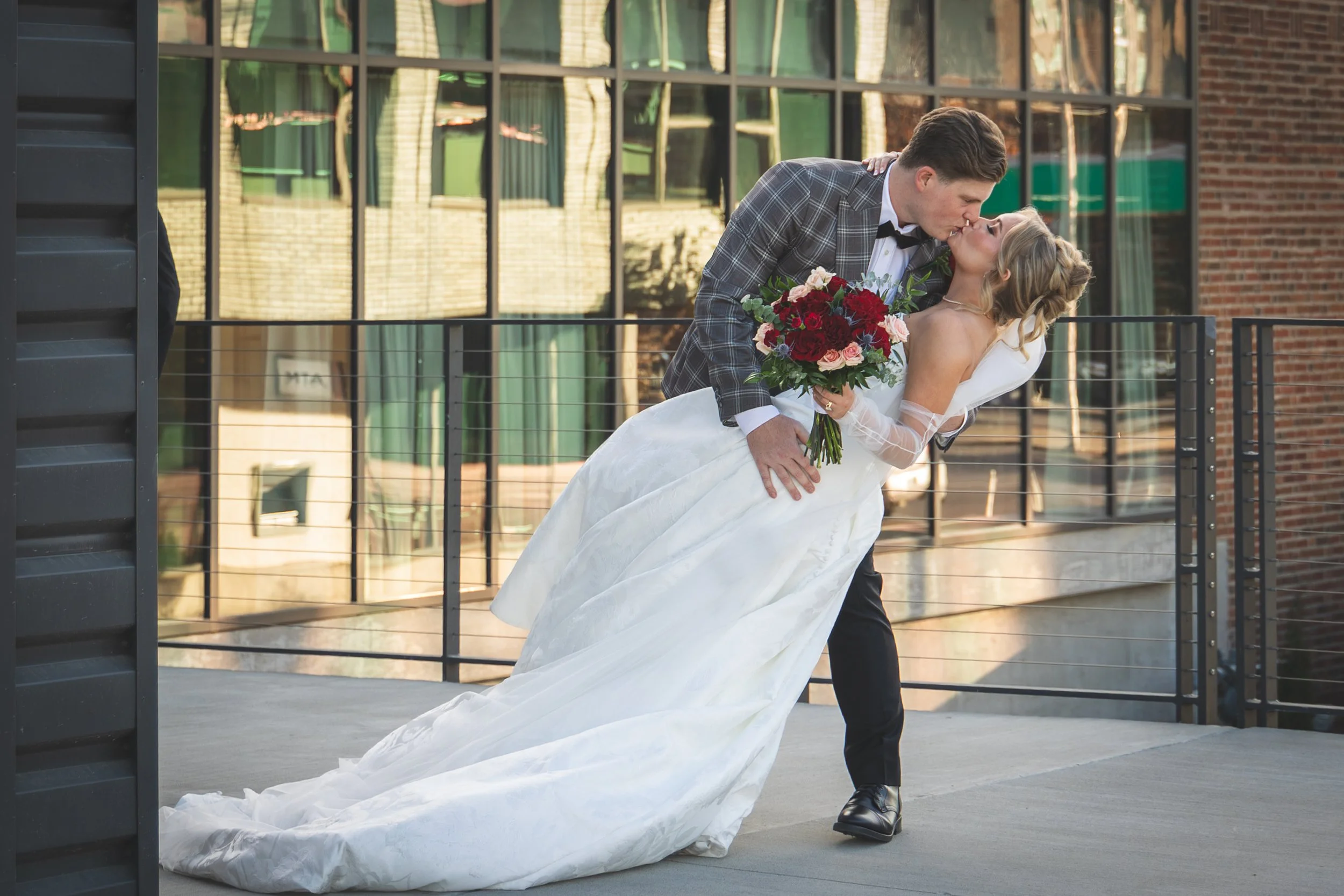 A groom in a checkered suit and bow tie dips and kisses a bride in a white wedding gown, who holds a large bouquet of red and pink roses, on a balcony with a cityscape background.