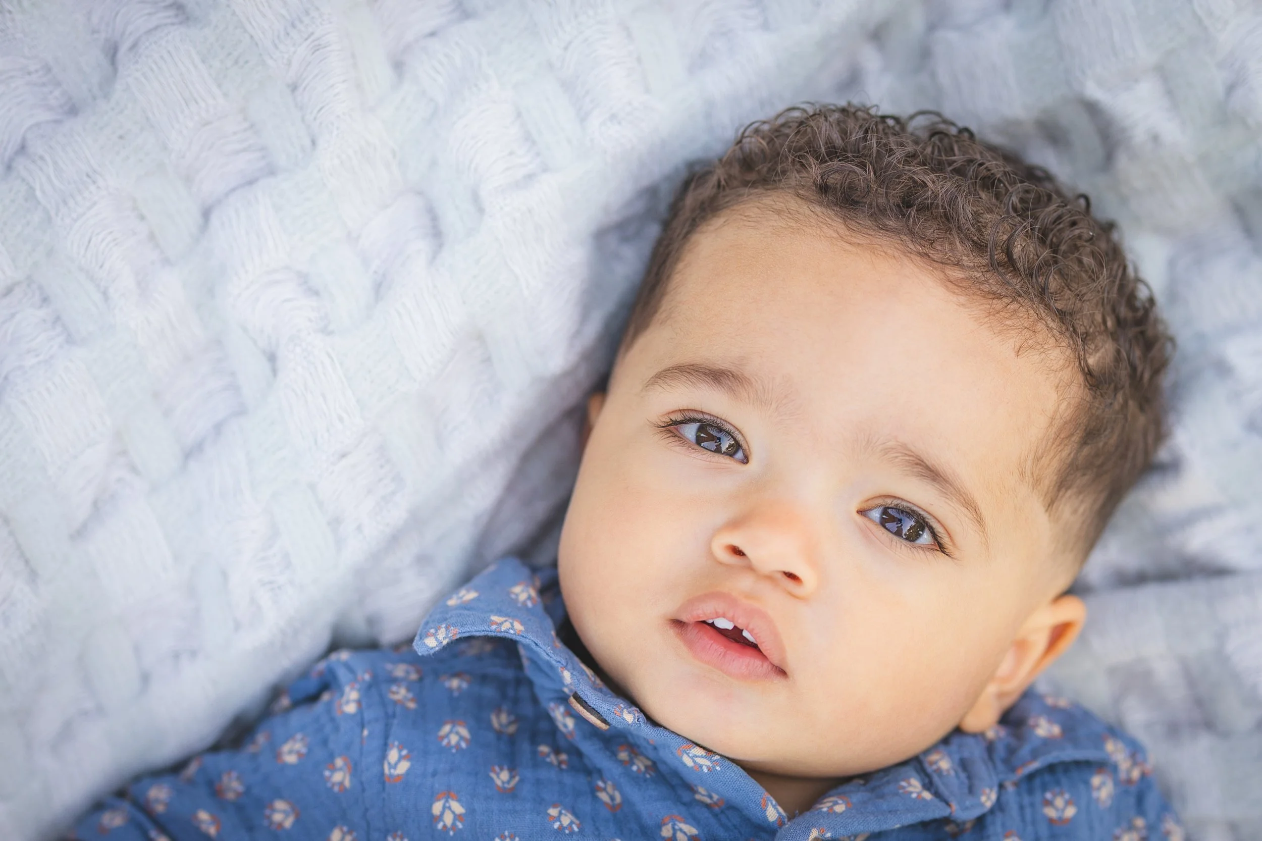 Close-up of a young child with curly hair lying on a textured white blanket, wearing a blue shirt with a floral pattern, looking at the camera.