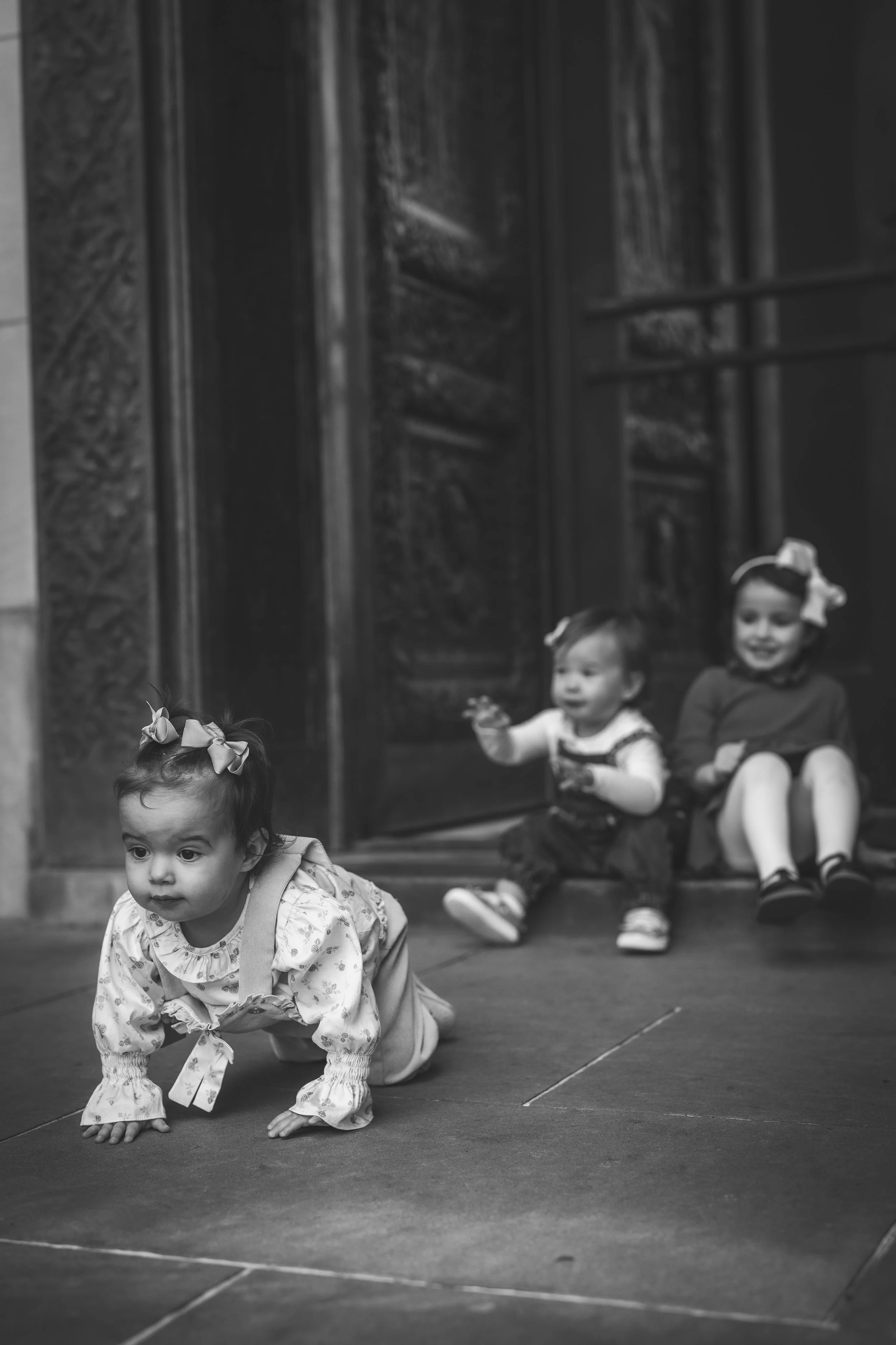 Three young girls sitting and playing on the floor in front of a wooden door, with one girl crawling forward in the foreground and two girls sitting behind.