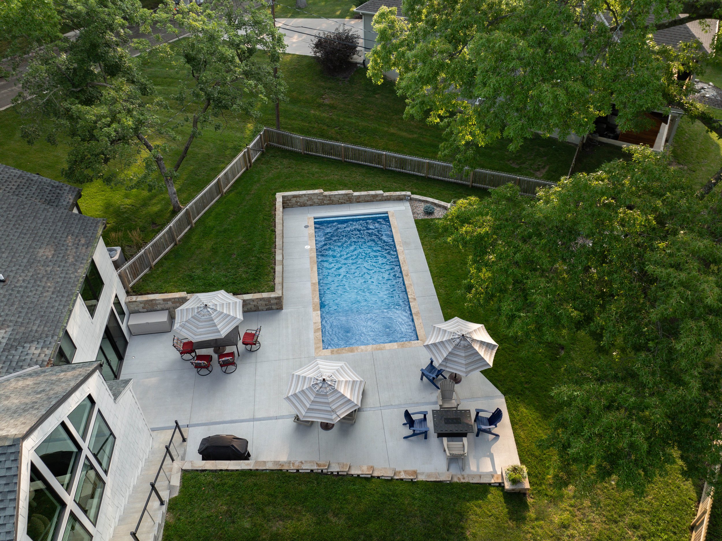 A backyard with a concrete patio, a rectangular pool, several patio umbrellas, chairs, and a grassy yard enclosed by a wooden fence. Tall trees with green leaves surround the area.