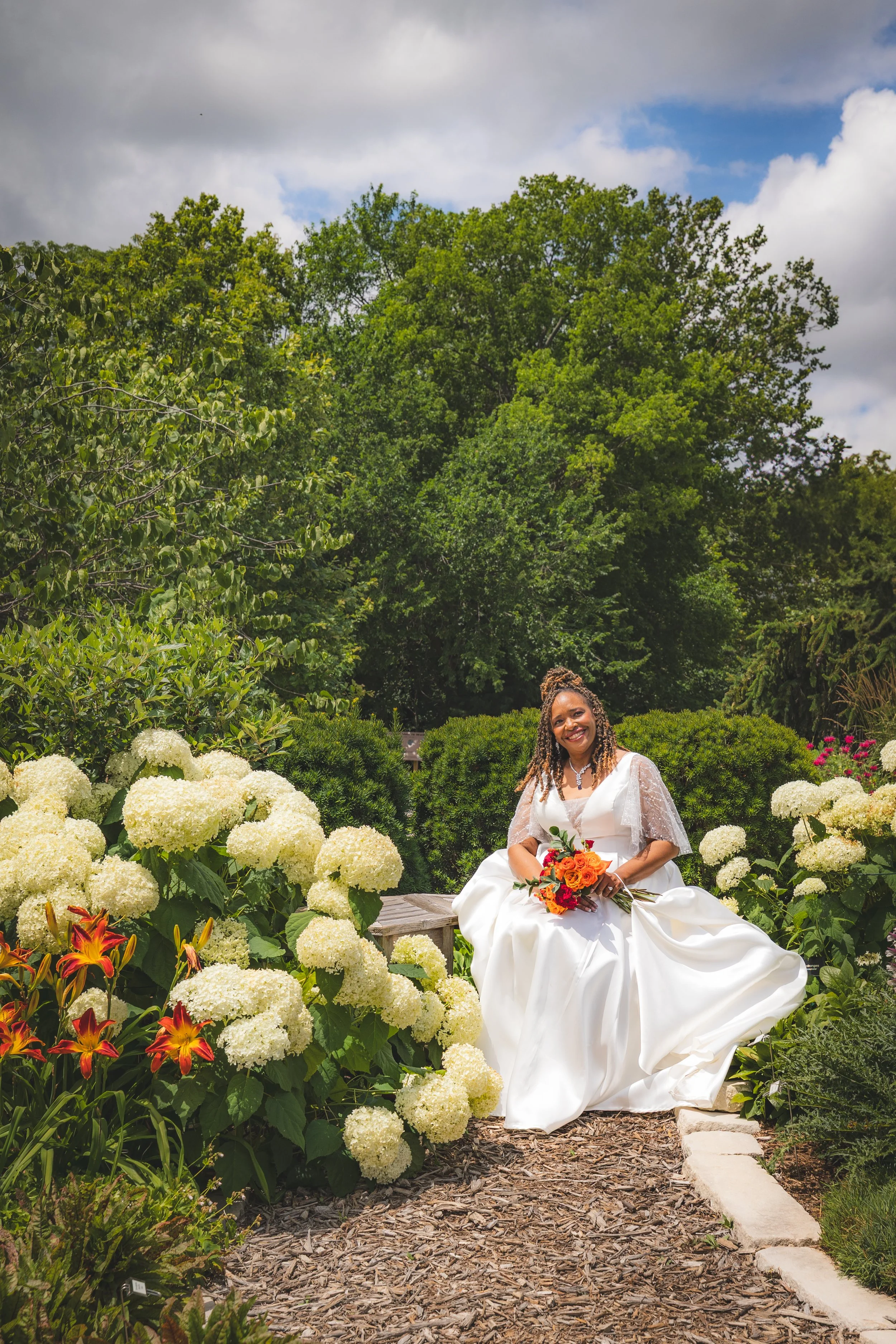 A woman in a white wedding dress sitting on a bench in a garden surrounded by white hydrangeas and orange lilies, holding a bouquet of orange and yellow flowers, smiling at the camera.