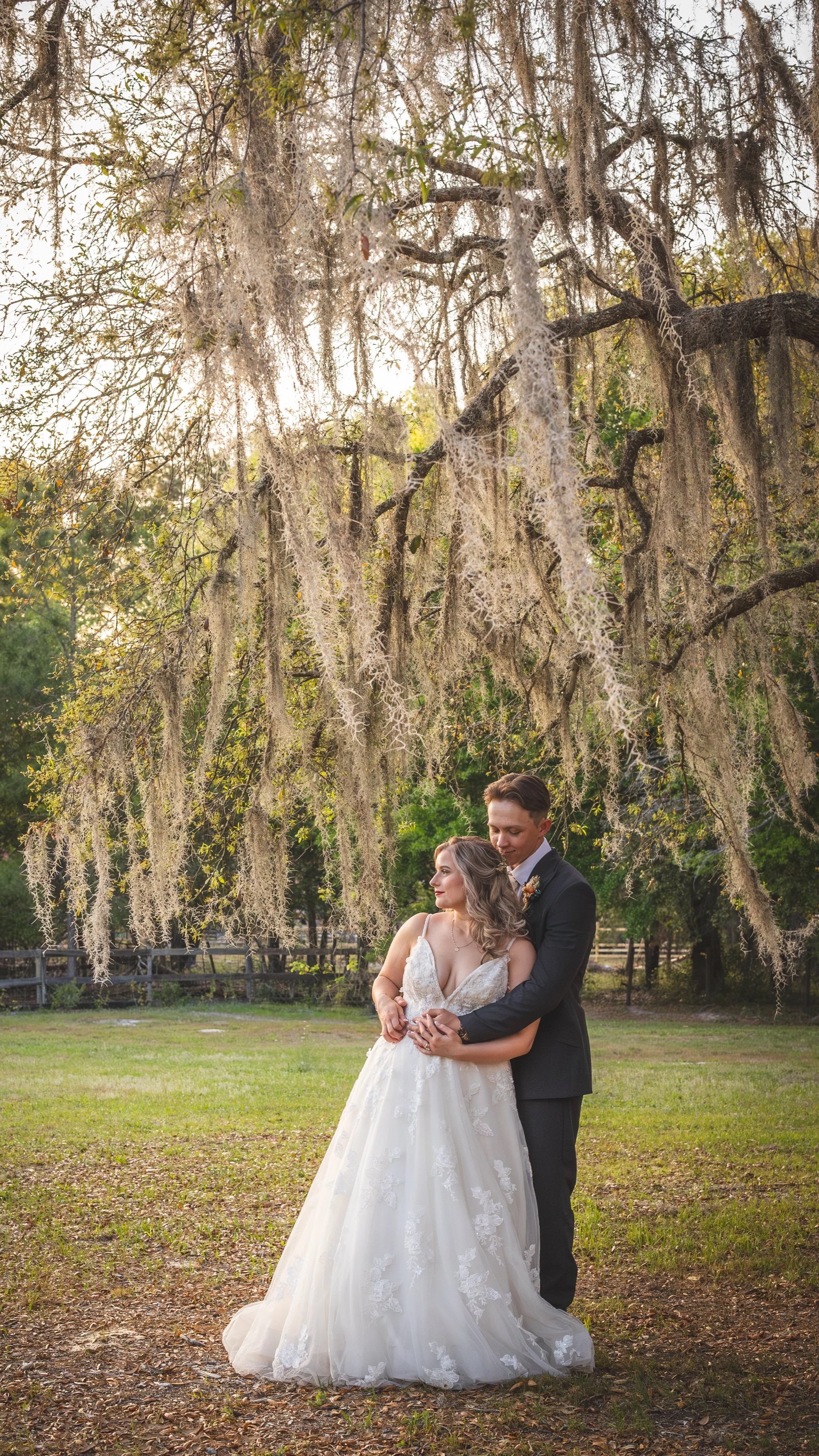 A bride and groom standing closely together under a large tree with hanging moss, in an outdoor setting during sunset.
