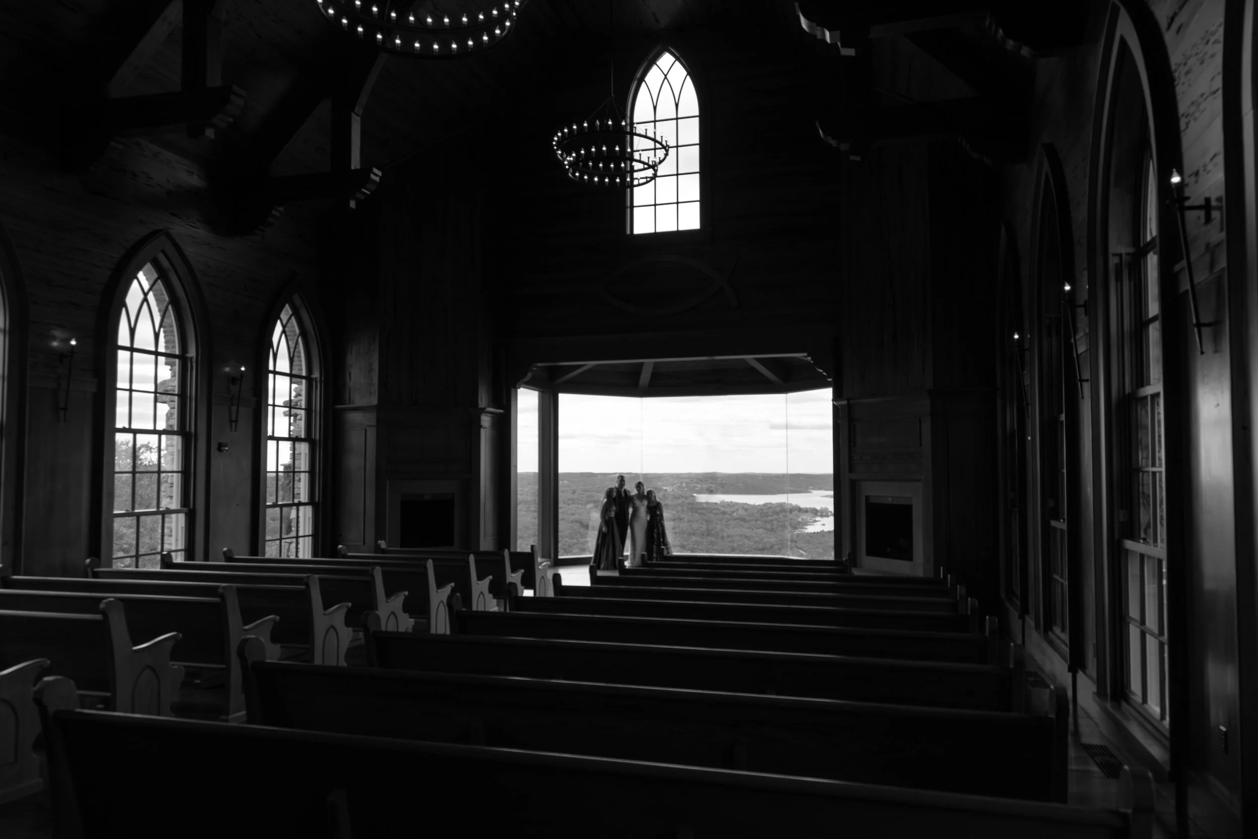 The interior of a church with large stained glass windows and wooden pews, showing a view of the outside landscape and three people standing near the front