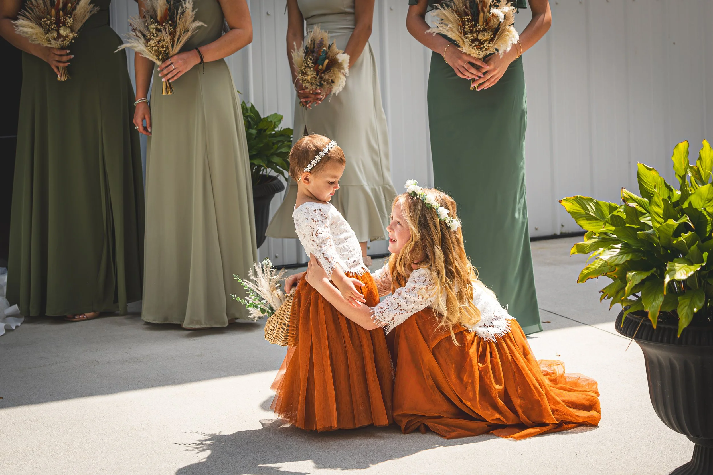 A young girl in a white lace top and orange skirt kneeling and embracing a younger girl in a matching white lace top and orange skirt, both wearing floral headbands, during a wedding ceremony with bridesmaids in green and beige dresses holding bouque
