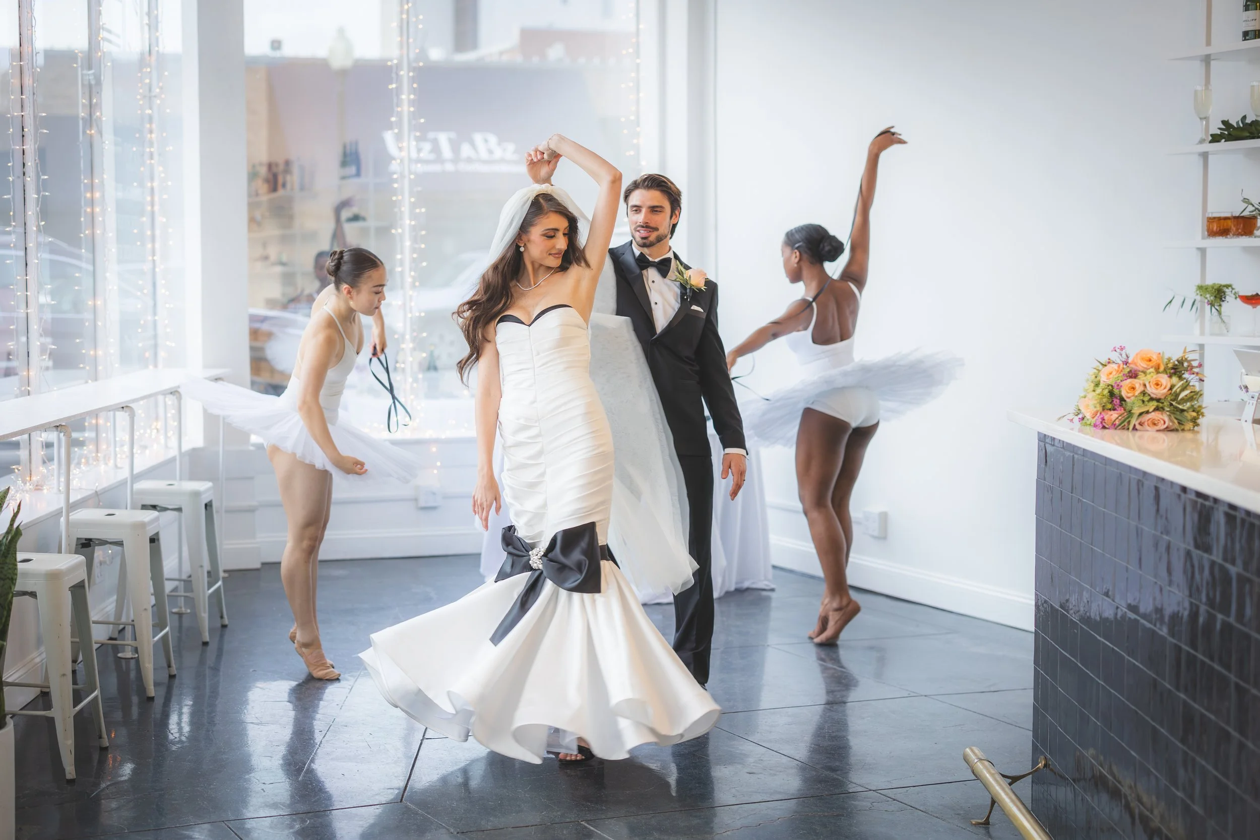 A bride and groom dancing in a brightly lit modern venue, with bridesmaids in tutus nearby and a bouquet on the counter.