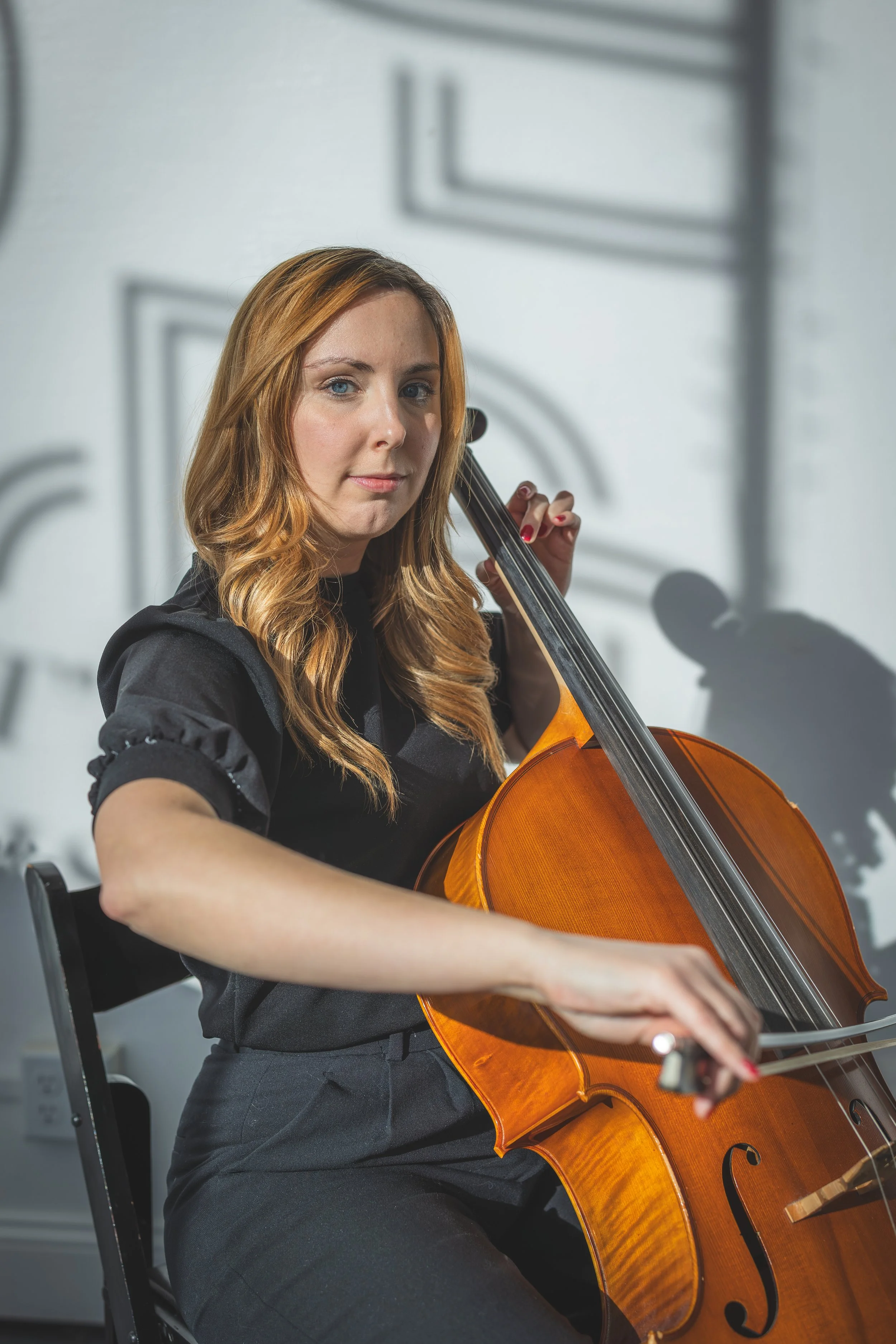 A woman with long, wavy red hair playing a cello in a room with plain walls and artistic wall decorations.