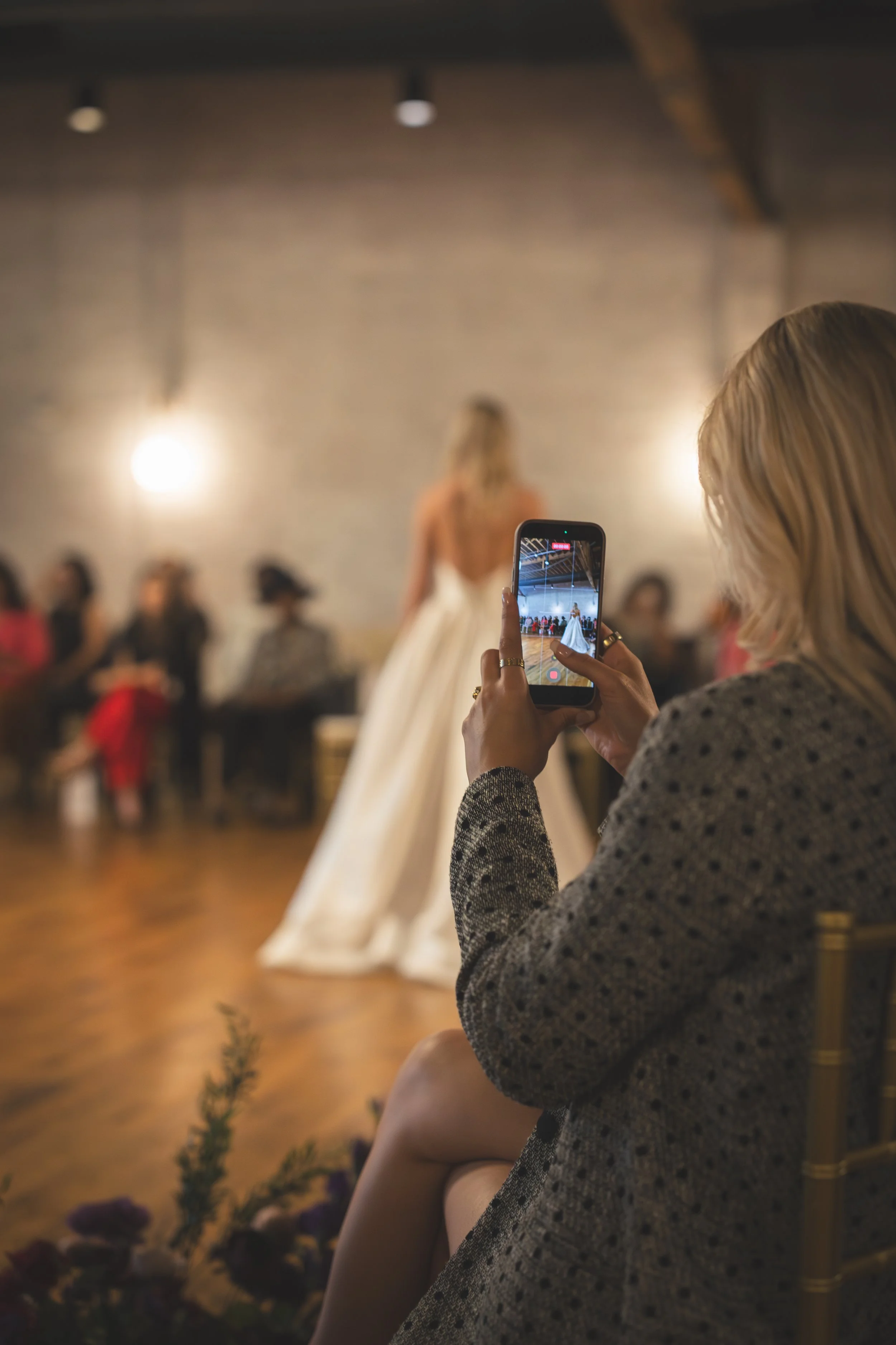 A woman with blonde hair taking a photo of a bride in a wedding dress with her phone at a wedding reception indoors.