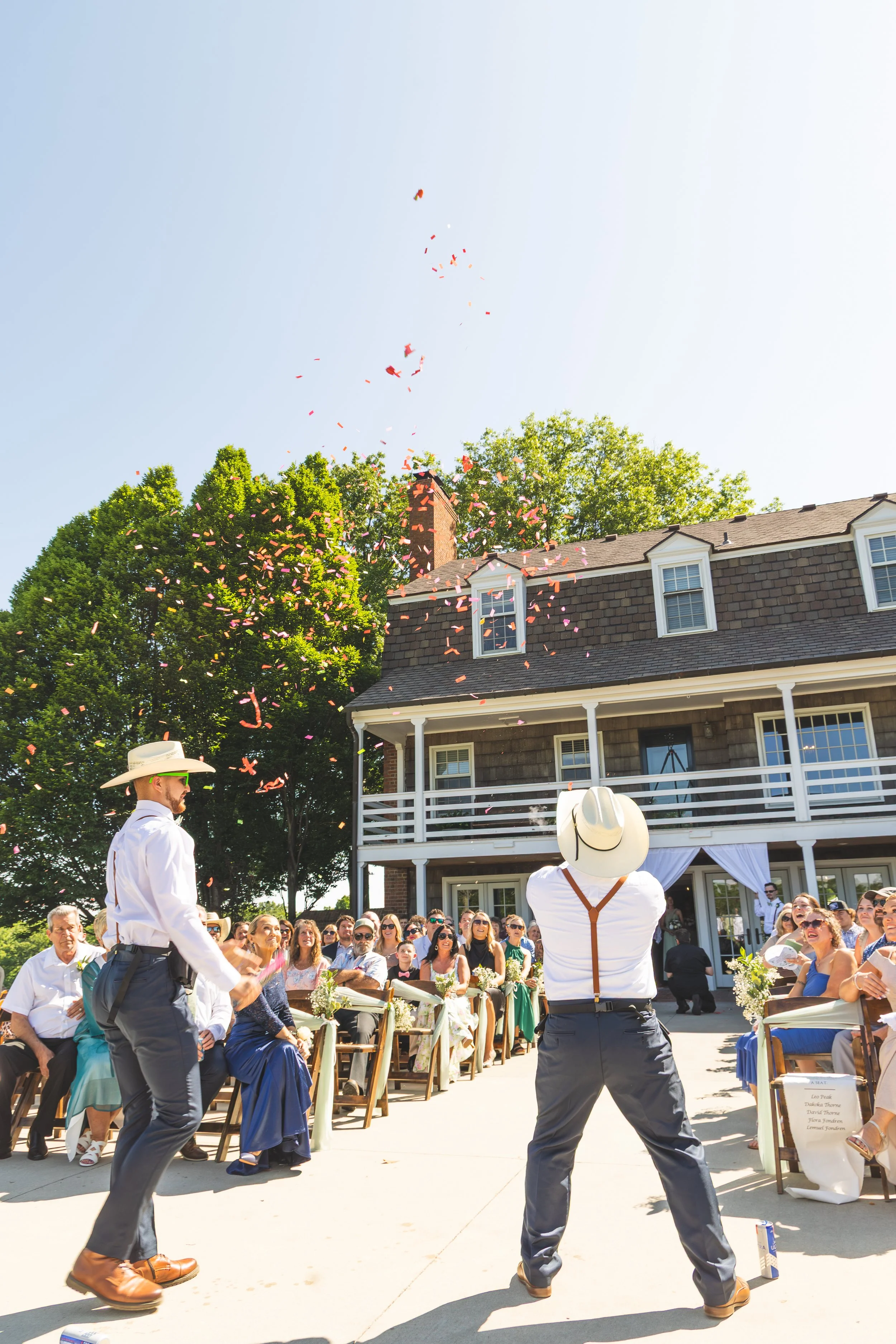 People attending a wedding ceremony outdoors with a focus on two men dressed in white shirts and gray pants, one with suspenders, standing in front of seated guests under a clear blue sky, with confetti falling from above and a house in the backgroun