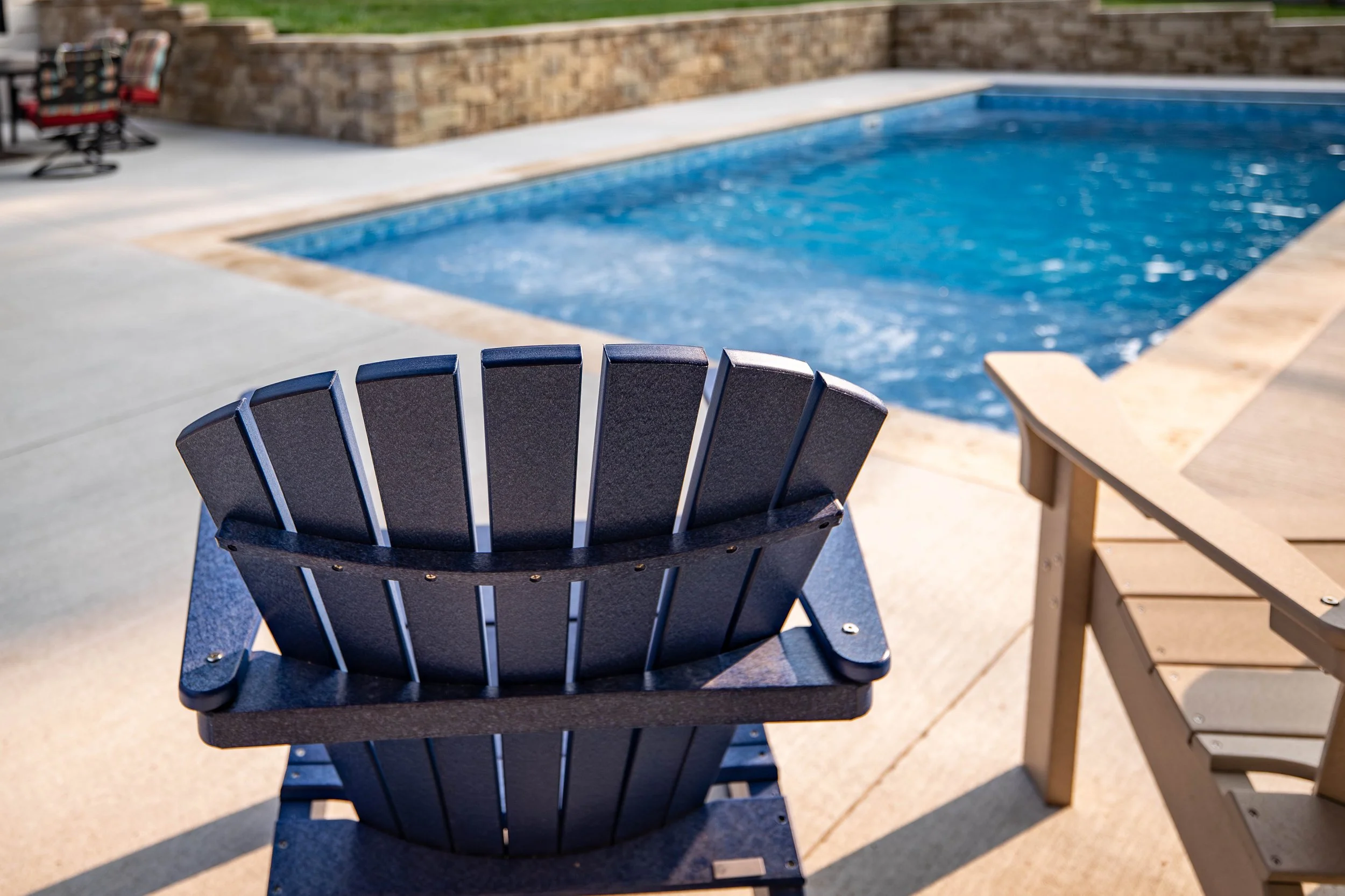 Black Adirondack chair on a concrete patio near a swimming pool with a brick wall and some outdoor furniture in the background.