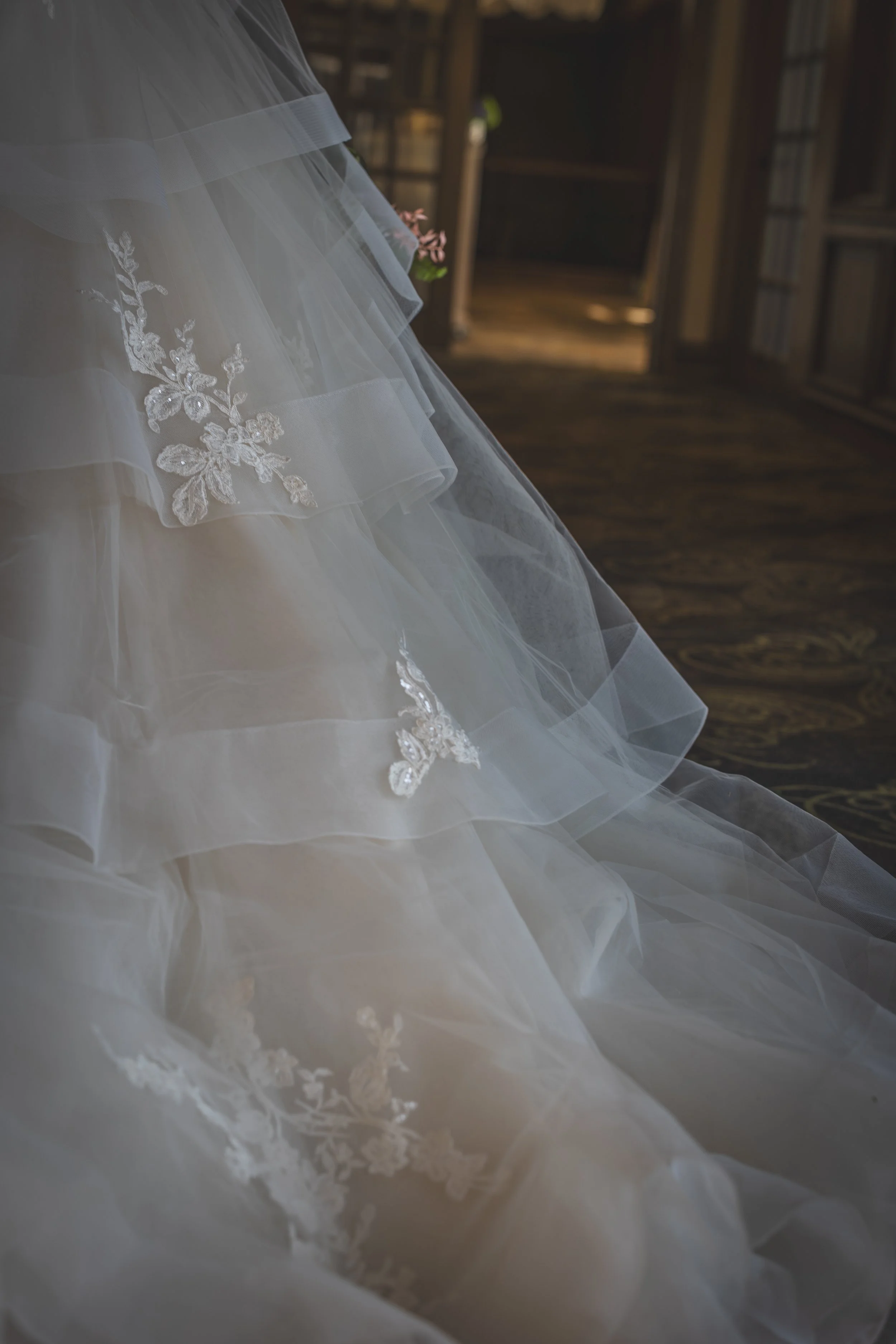 Close-up of a wedding dress with lace embroidery and layered tulle fabric, hanging indoors with a wooden bookshelf in the background.