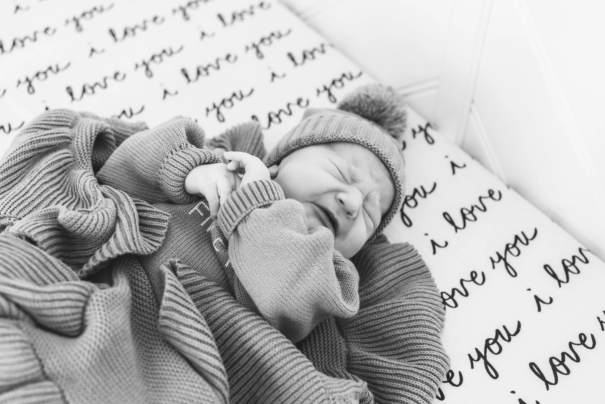 A newborn baby wearing a knit hat and sweater, lying on a blanket with handwritten words 'love you' repeatedly.